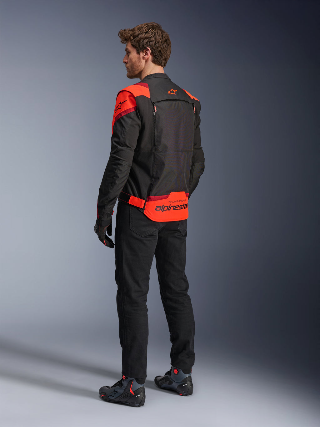 Alpinestars T-Stunt Air Jacket van achteren gezien, zwart, fluorood en donkerrood, textiel motorjas met mesh-panelen en sportieve snit, met opvallende Alpinestars-branding op de onderrug, man draagt de jas met een zwarte broek en motorlaarzen