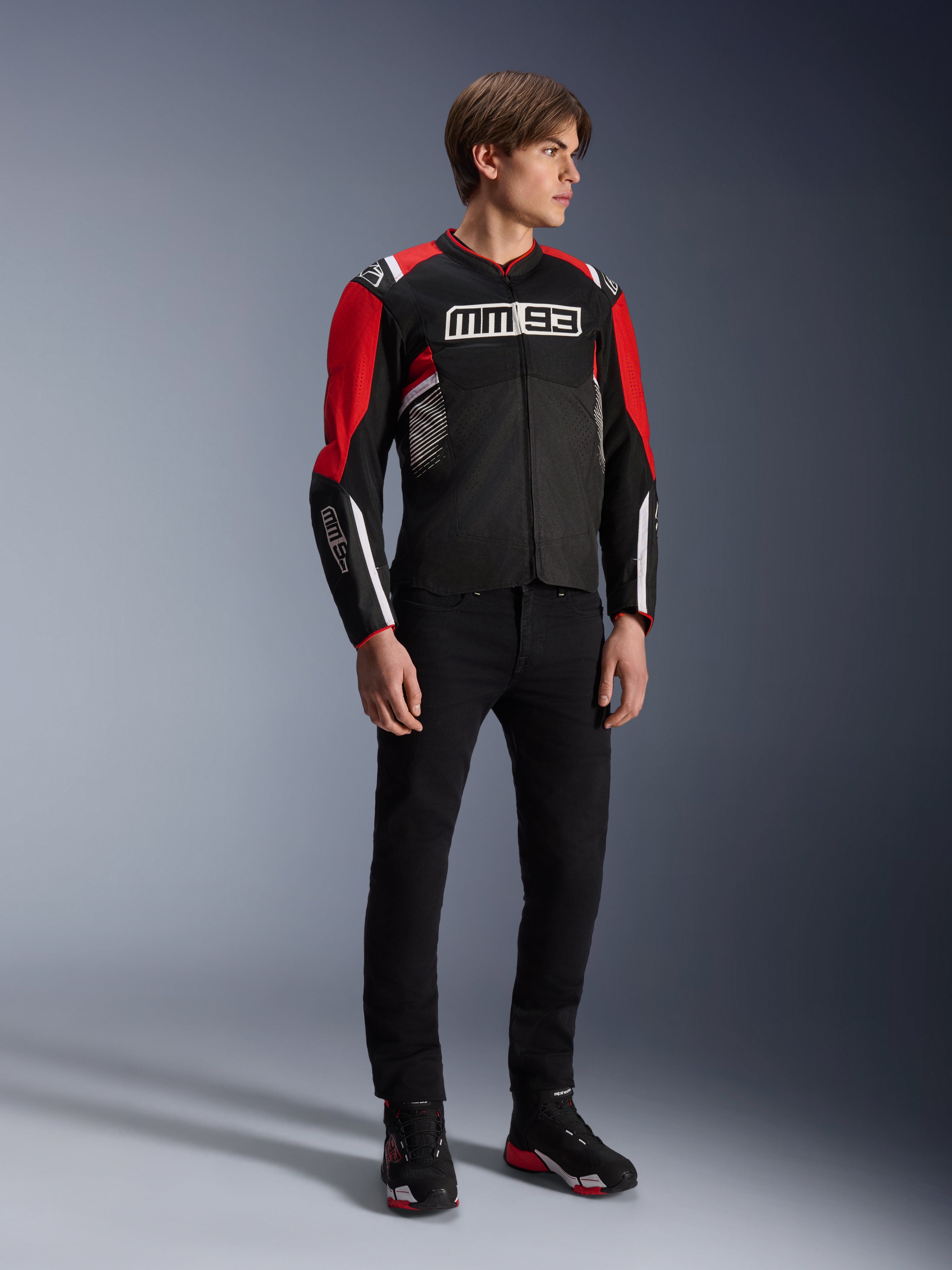 Mannelijk model draagt de Alpinestars MM93 T-SPS Air v2 Jas in Black Bright Red White, met opvallende MM93-branding op de borst en mouwen, gecombineerd met een zwarte broek en motorlaarzen, sportieve urban motorstijl met zichtbare mesh-panelen voor luchtstroom.