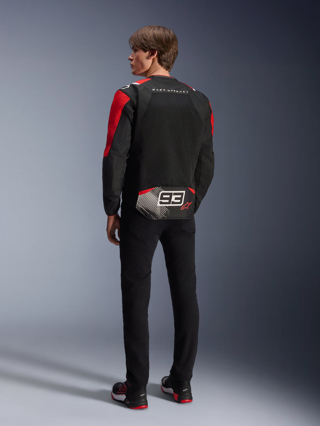Model van achteren gezien met de Alpinestars MM93 T-SPS Air V2 Jas, zwart met felrode en witte details, voorzien van 'MARC MARQUEZ' tekst en een groot '93' logo op de onderrug, mesh textielconstructie voor ventilatie, gedragen met een zwarte broek en bijpassende zwart-rode motorschoenen