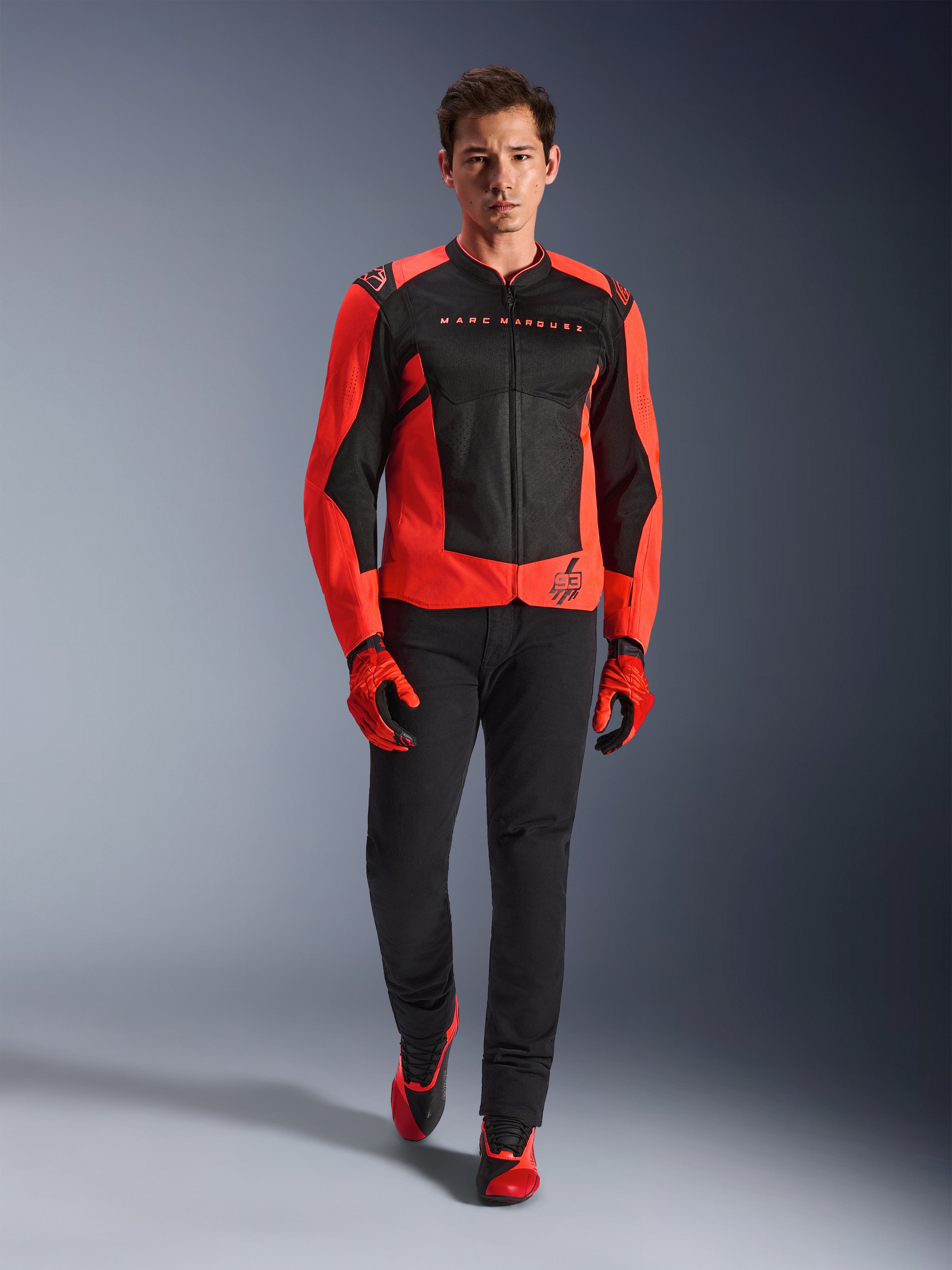 Alpinestars MM93 T-SPS Air V2 Jas, fluo rood en zwart, gedragen door een model met bijpassende rode handschoenen en zwarte broek, voorzien van Marc Marquez-branding en het 93-logo, geventileerde sportieve motorjas voor stadsritten