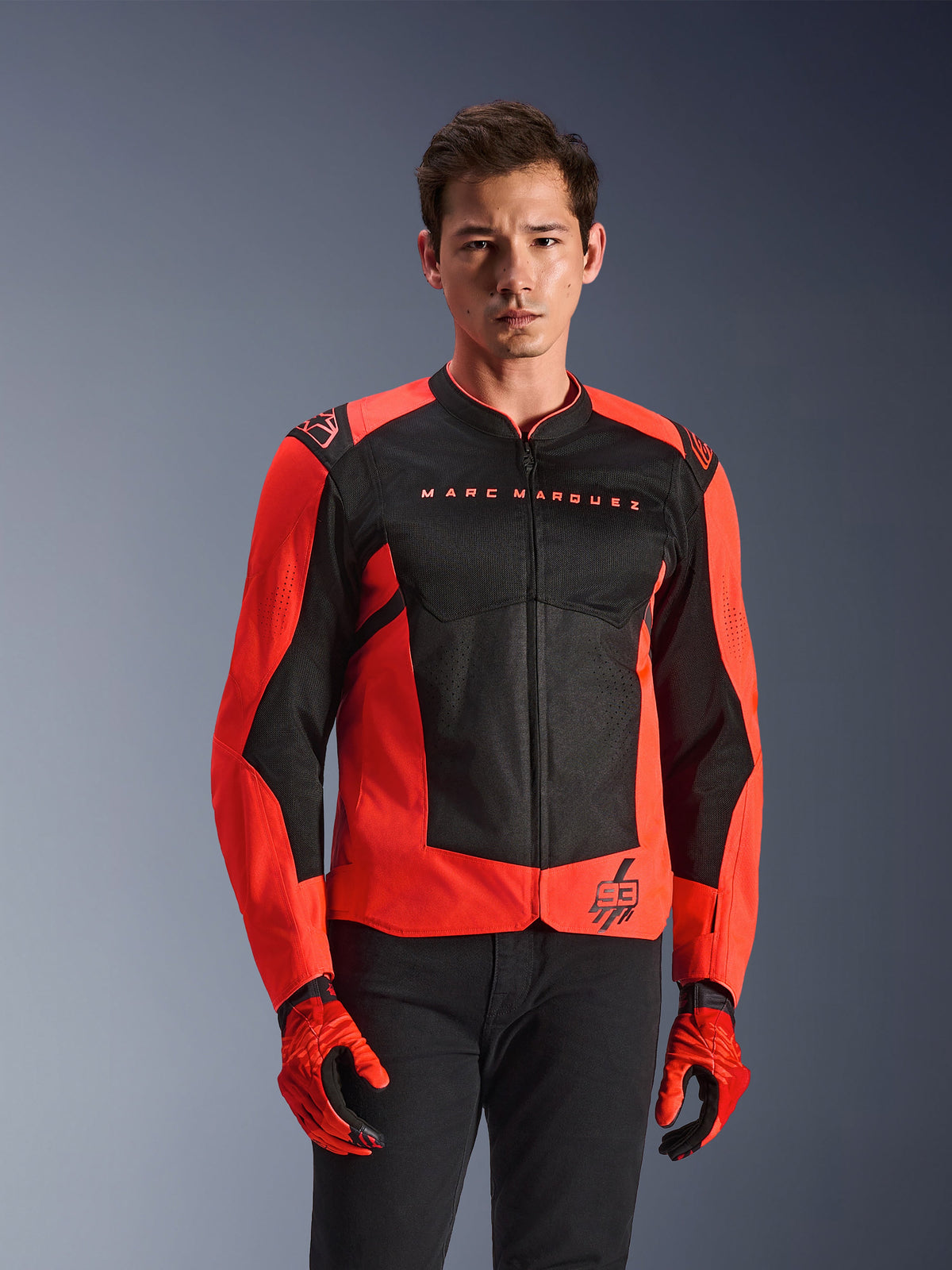 Veste Alpinestars MM93 T-SPS Air V2, veste de moto, rouge fluo et noir, portée par un mannequin avec des gants rouges assortis et un pantalon noir, logo 'Marc Marquez' sur la poitrine et logo '93', construction sportive en mesh