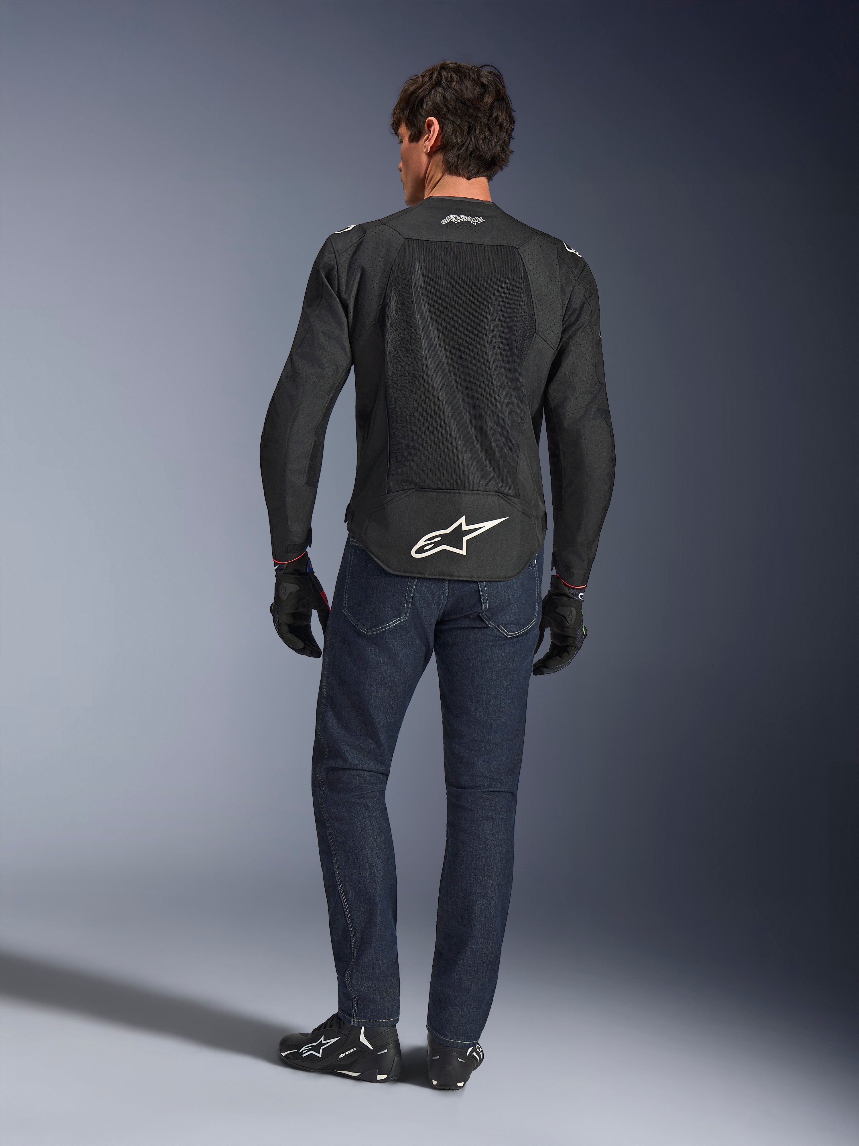 Alpinestars FQ20 T-SPS Air V2 Monster Jas, zwart, achteraanzicht op model met donkerblauwe jeans en zwarte motorlaarzen, voorzien van een groot wit logo op de onderrug en ademende mesh-panelen, stedelijke sportieve motoruitrusting.