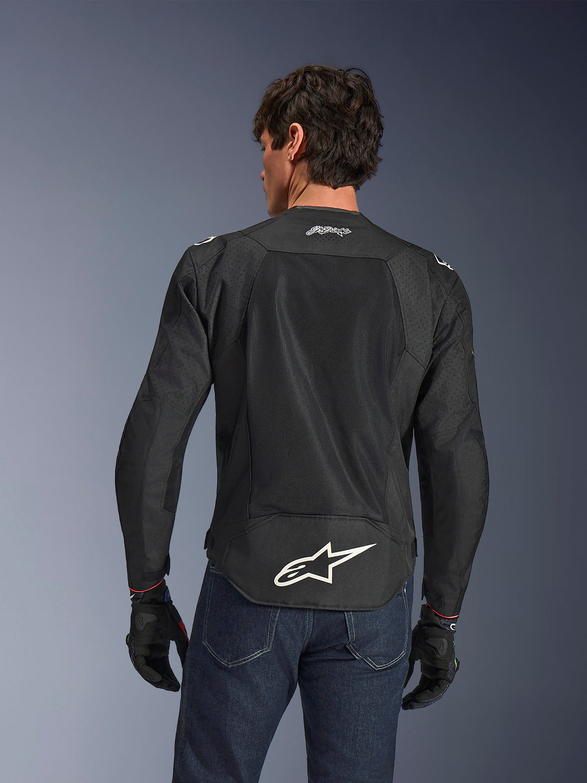 Alpinestars FQ20 T-SPS Air V2 Monster Jas, zwart, achteraanzicht met grote ventilatie-meshpanelen en wit Alpinestars-logo op de onderrug, gedragen met blauwe jeans en zwarte motorhandschoenen, stedelijke sportieve rijstijl.