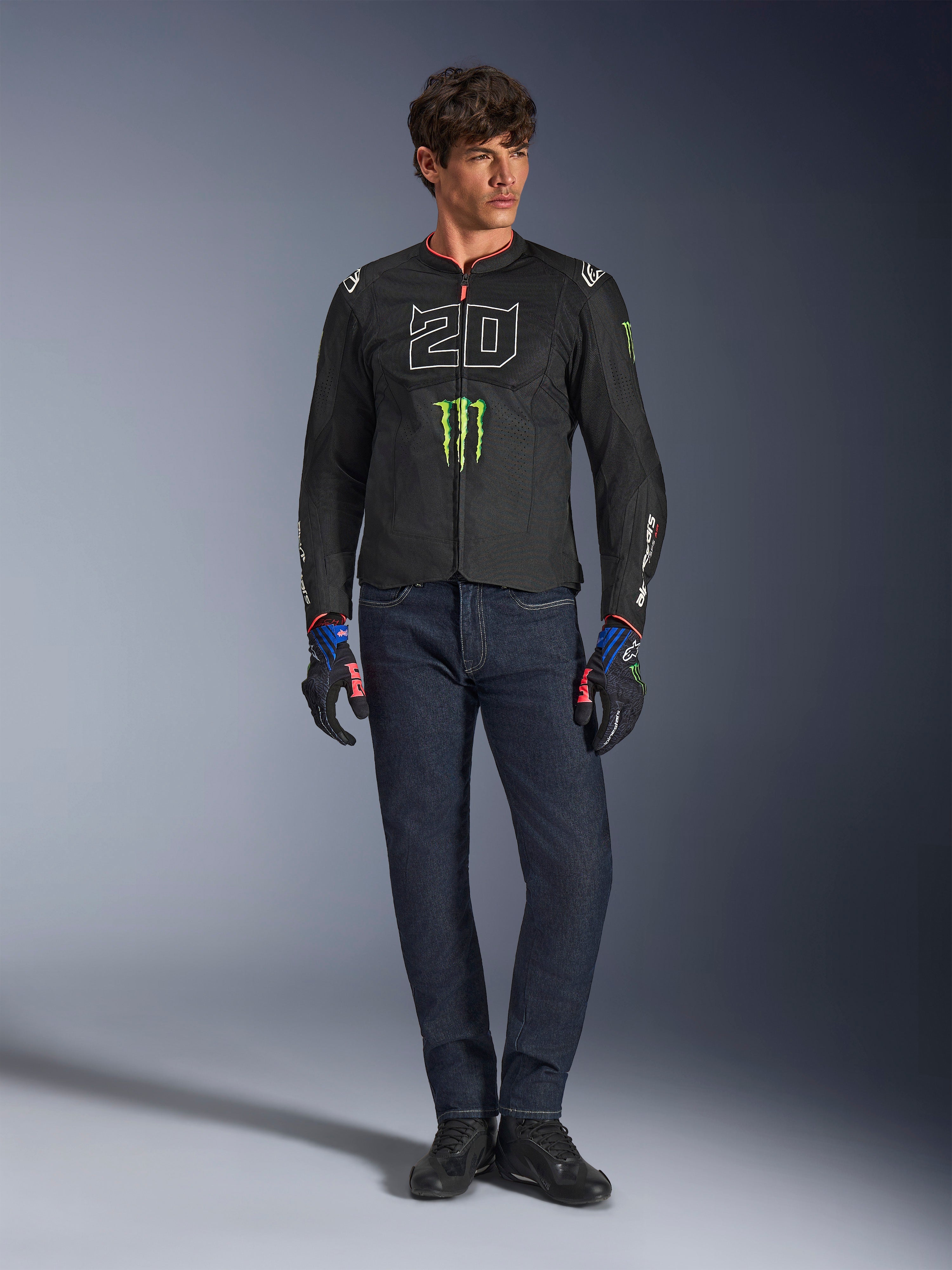 Mannelijke rijder die de Alpinestars FQ20 T-SPS Air V2 Monster jas draagt, zwart met groen Monster Energy-logo en witte '20' graphic op de borst, gecombineerd met een donkerblauwe jeans en technische motorhandschoenen, urban sportieve motoroutfit.