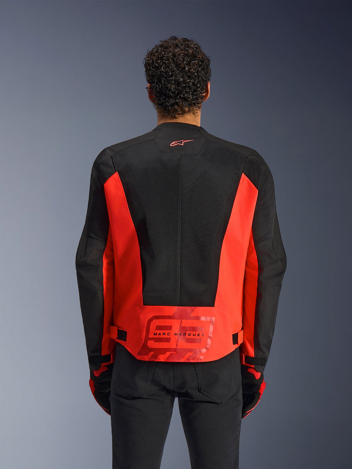 Achteraanzicht van een man die de Alpinestars MM93 C-1 Air Jacket draagt in Red Fluo en zwart, voorzien van een groot '93' Marc Marquez-logo op de onderrug en lichtgewicht mesh-panelen voor ventilatie, gecombineerd met een zwarte broek en handschoenen.