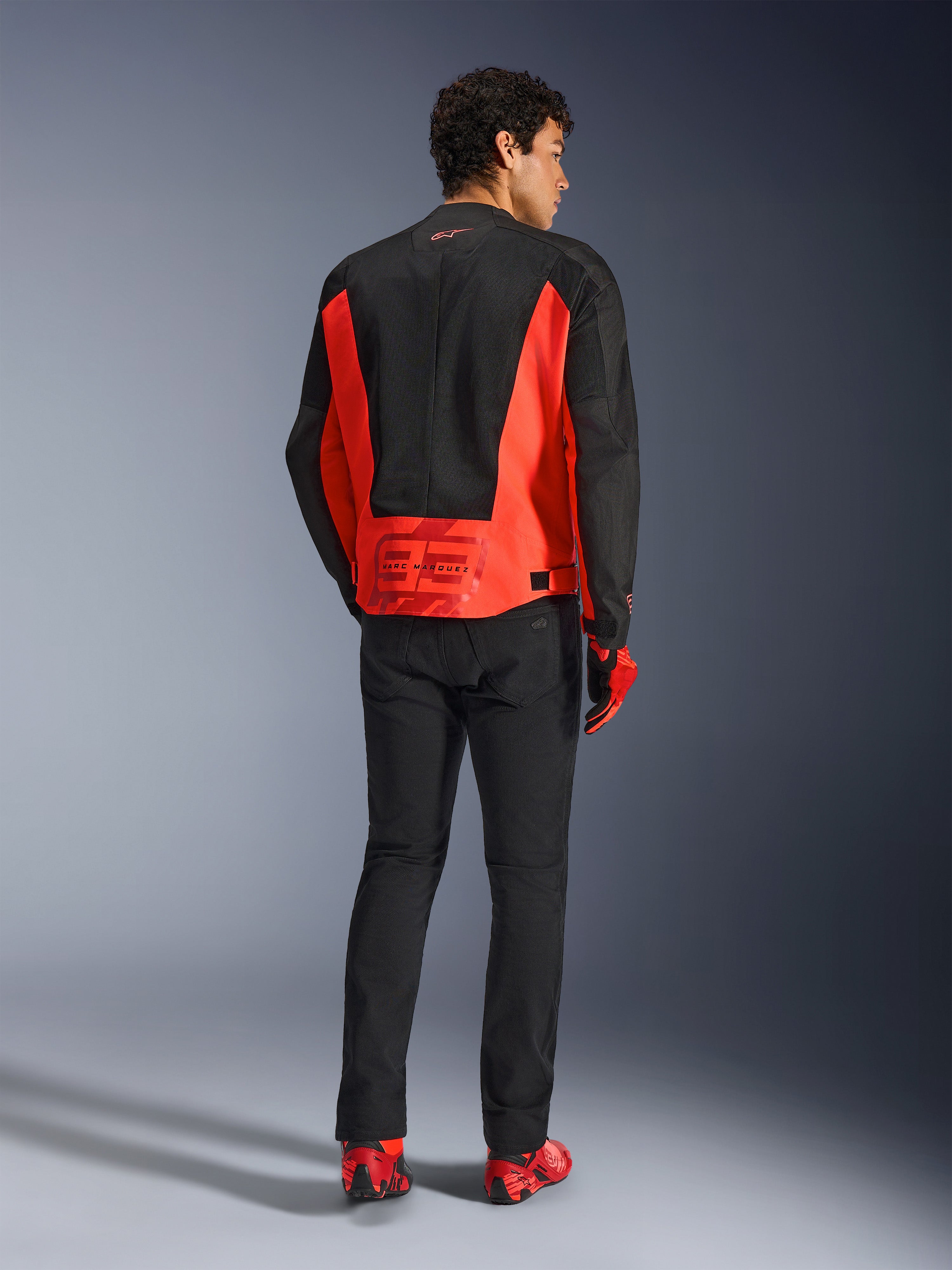Achteraanzicht van een mannelijk model dat de Alpinestars MM93 C-1 Air Jas in fluo rood en zwart draagt, voorzien van het Marc Marquez 93-logo op de onderrug, gecombineerd met een zwarte broek en rode motorschoenen, stedelijke motor woon-werkstijl