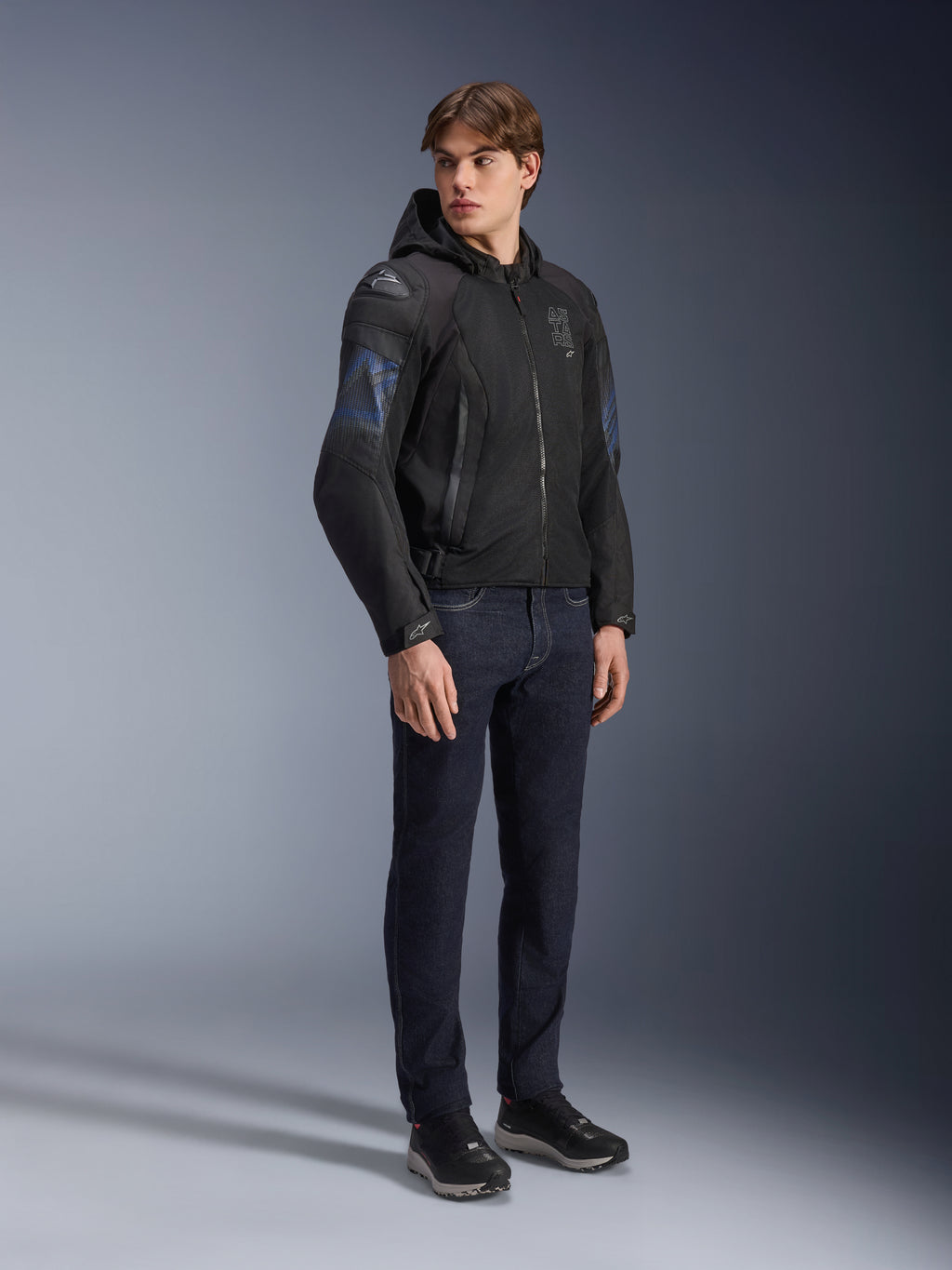 Alpinestars Zaca Air Venom WP Jacke, Textil-Motorradjacke in Schwarz und Electric Blue, getragen von einem Model mit dunkler Jeans und schwarzen Sneakern, mit abnehmbarer Kapuze, Mesh-Belüftungseinsätzen und DFS Lite Schulterschleifern, sportliche Allwetter-Fahrausrüstung.