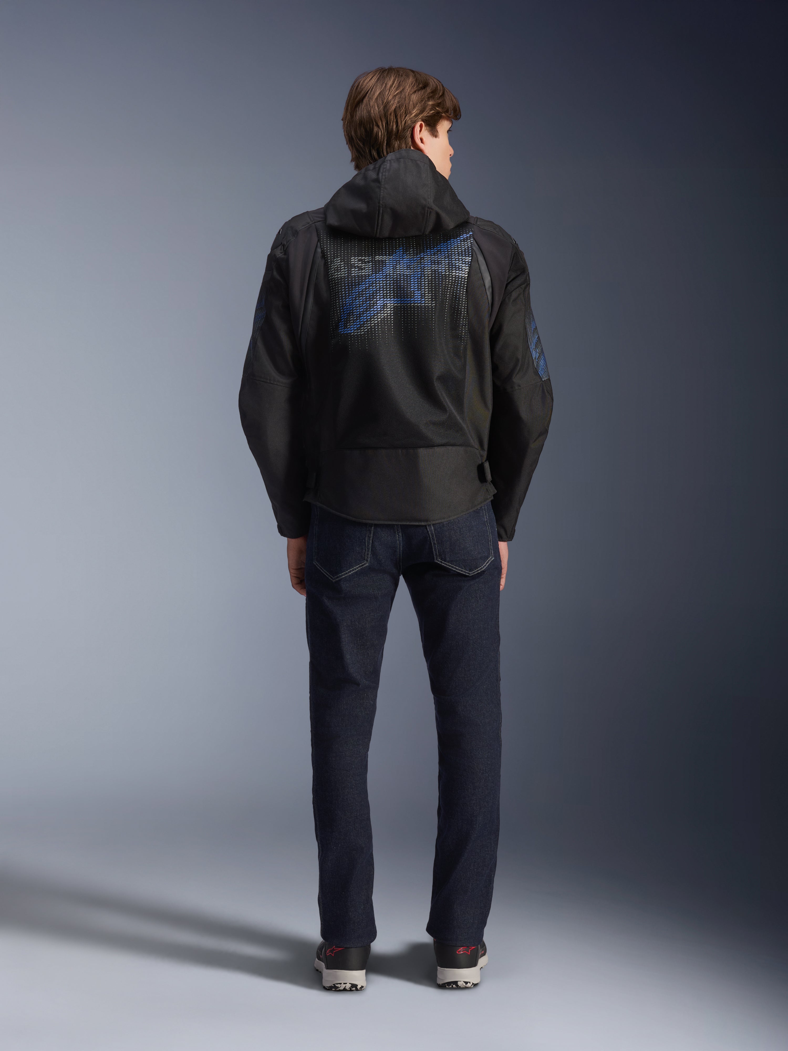 Alpinestars Zaca Air Venom WP Jacke, Schwarz und Electric Blue, Rückansicht mit einem großen blauen Alpinestars-Logo auf dem Mesh-Rückeneinsatz, abnehmbare Kapuze, getragen mit dunklen Denim-Jeans und Sneakern, sportliche Allwetter-Motorradausrüstung.