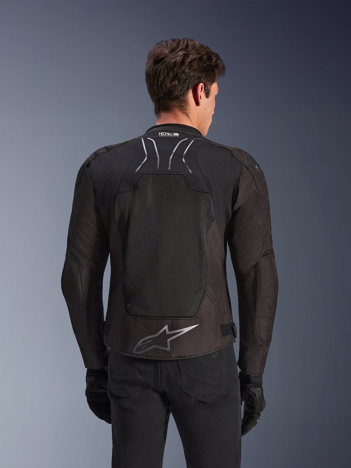 Veste Alpinestars SMX Air en Black Anthracite, vue de dos sur un mannequin, dotée d'un grand panneau dorsal en maille technique pour la ventilation, marquage Tech-Air Ready sur le col et logo Astar argenté dans le bas du dos, portée avec des gants noirs et un pantalon de moto.