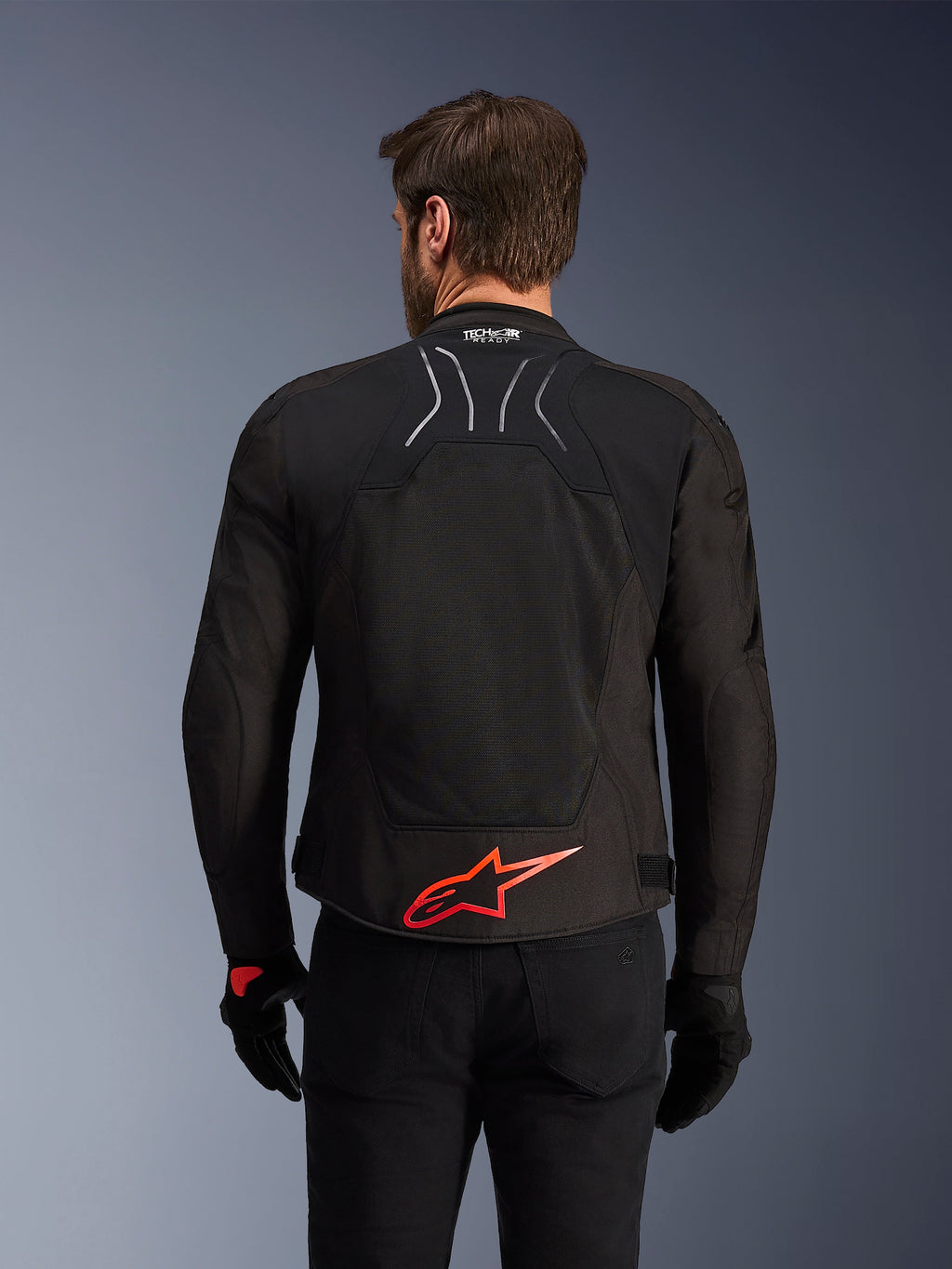 Veste Alpinestars SMX Air, vue arrière, noire avec logo Alpinestars rouge vif dans le bas du dos, dotée d'un panneau en maille technique à haute circulation d'air et du marquage Tech-Air Ready, portée par un pilote avec pantalon et gants noirs