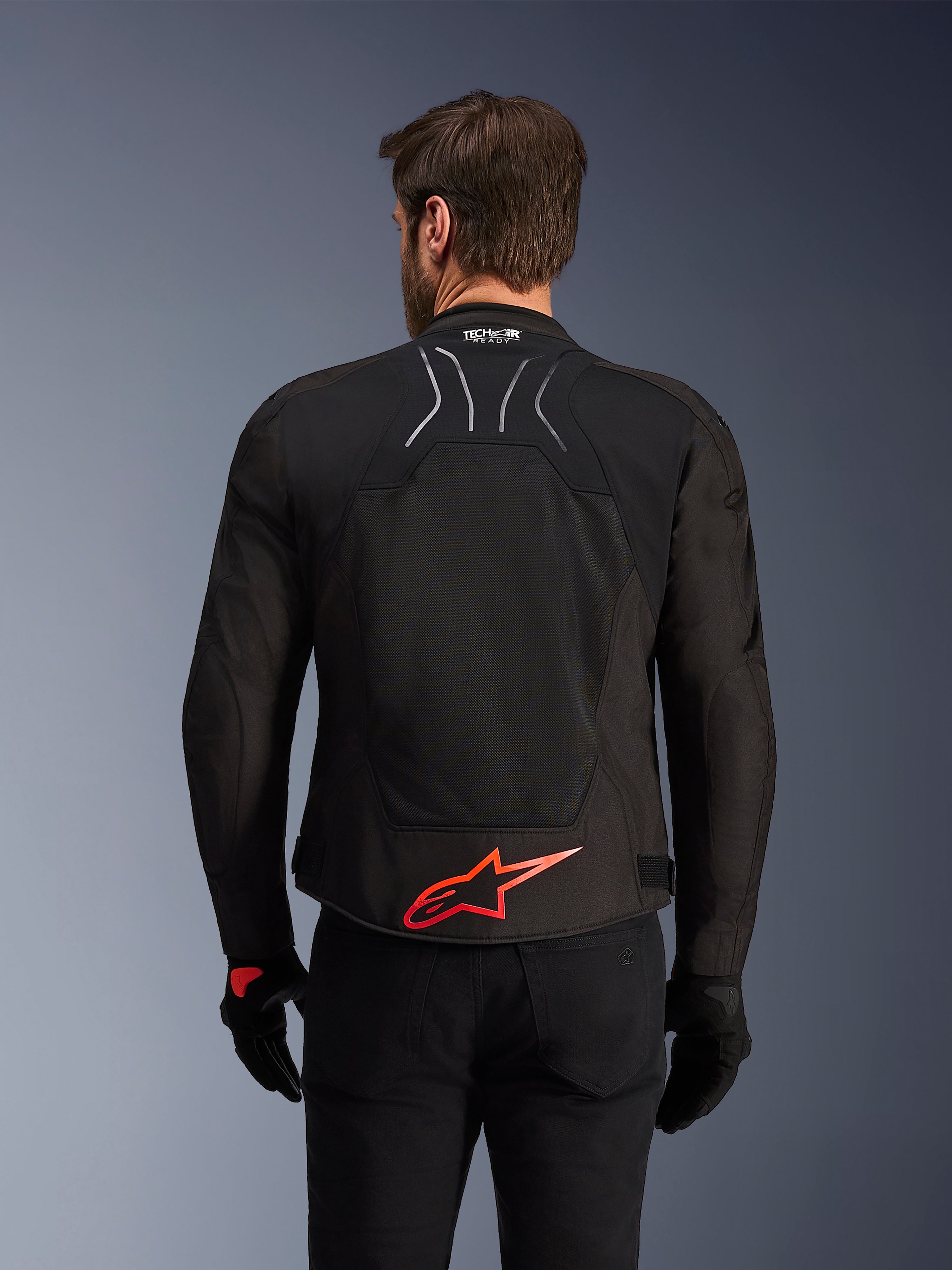 Veste Alpinestars SMX Air, vue arrière, noire avec logo Alpinestars rouge vif dans le bas du dos, dotée d'un panneau en maille technique à haute circulation d'air et du marquage Tech-Air Ready, portée par un pilote avec pantalon et gants noirs