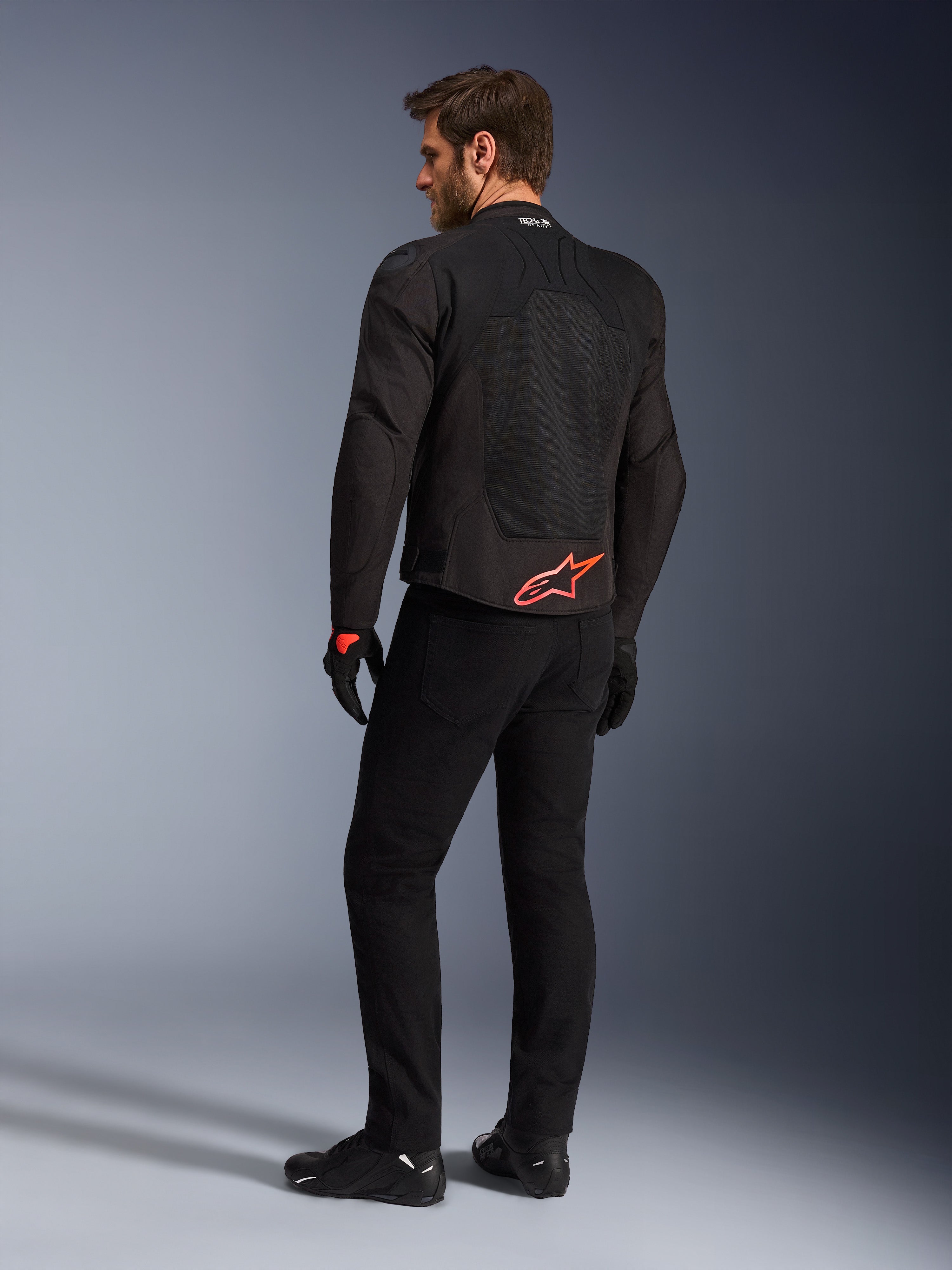 Vue arrière d'un mannequin portant la veste Alpinestars SMX Air en noir, arborant un logo Alpinestars rouge vif en bas du dos, le marquage « Tech-Air Ready » sur le col et des panneaux en maille technique pour la ventilation, associée à un pantalon et des gants de moto noirs.