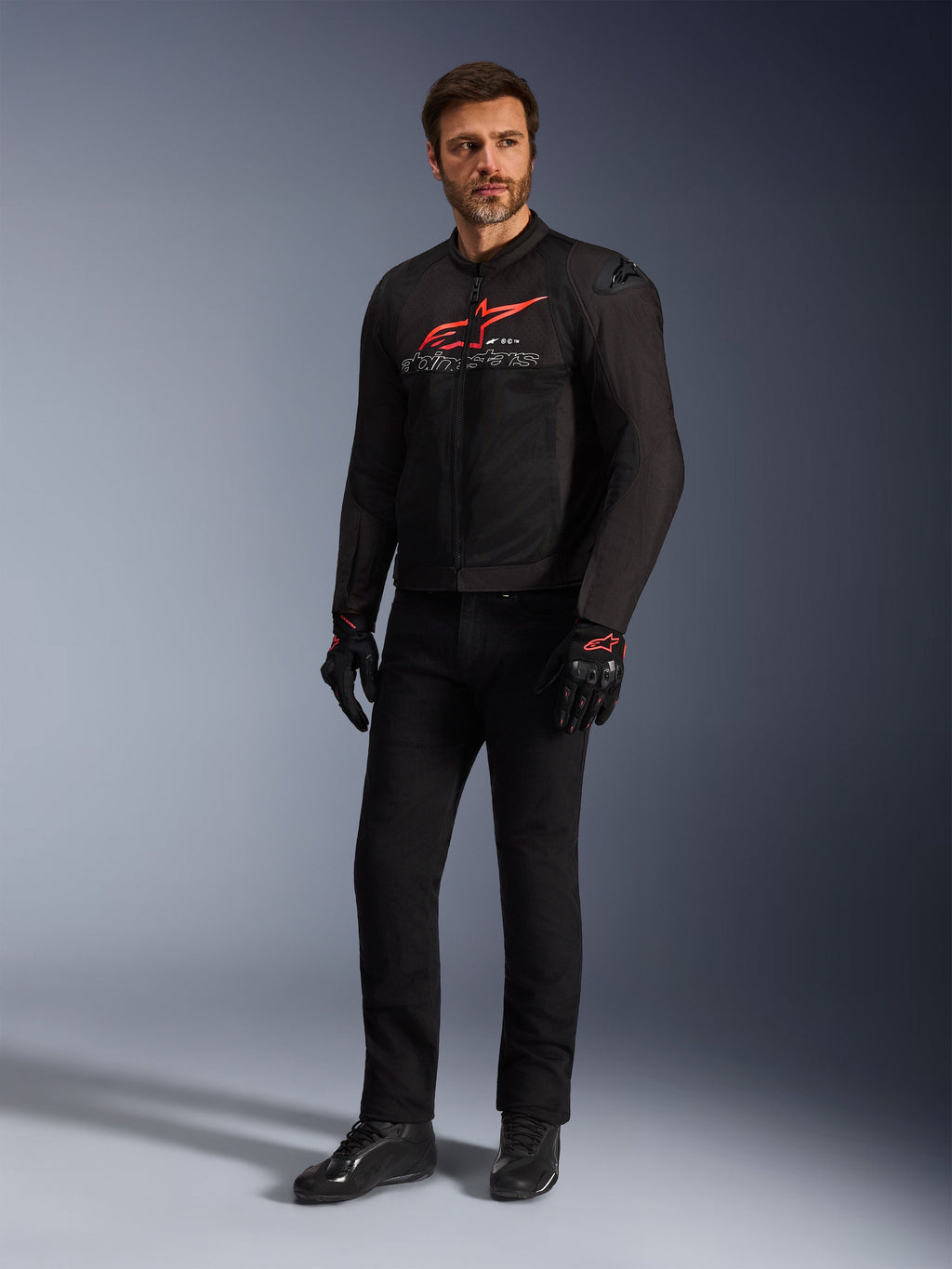Modèle masculin portant la veste Alpinestars SMX Air en noir, rouge vif et blanc, avec un pantalon technique noir, des gants noirs et rouges et des chaussures de moto noires, logo rouge distinctif sur la poitrine, photo de studio en pied pour équipement de moto sportive