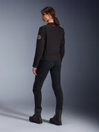 Model in der Alpinestars Stella Grace Jacke in Schwarz, Rückansicht mit Fokus auf den Vintage-Oscar-Spiralaufnäher am Arm und das kleine Stern-Logo am Kragen, kombiniert mit schwarzer Motorradhose und schwarzen Stiefeln für urbanes Fahren