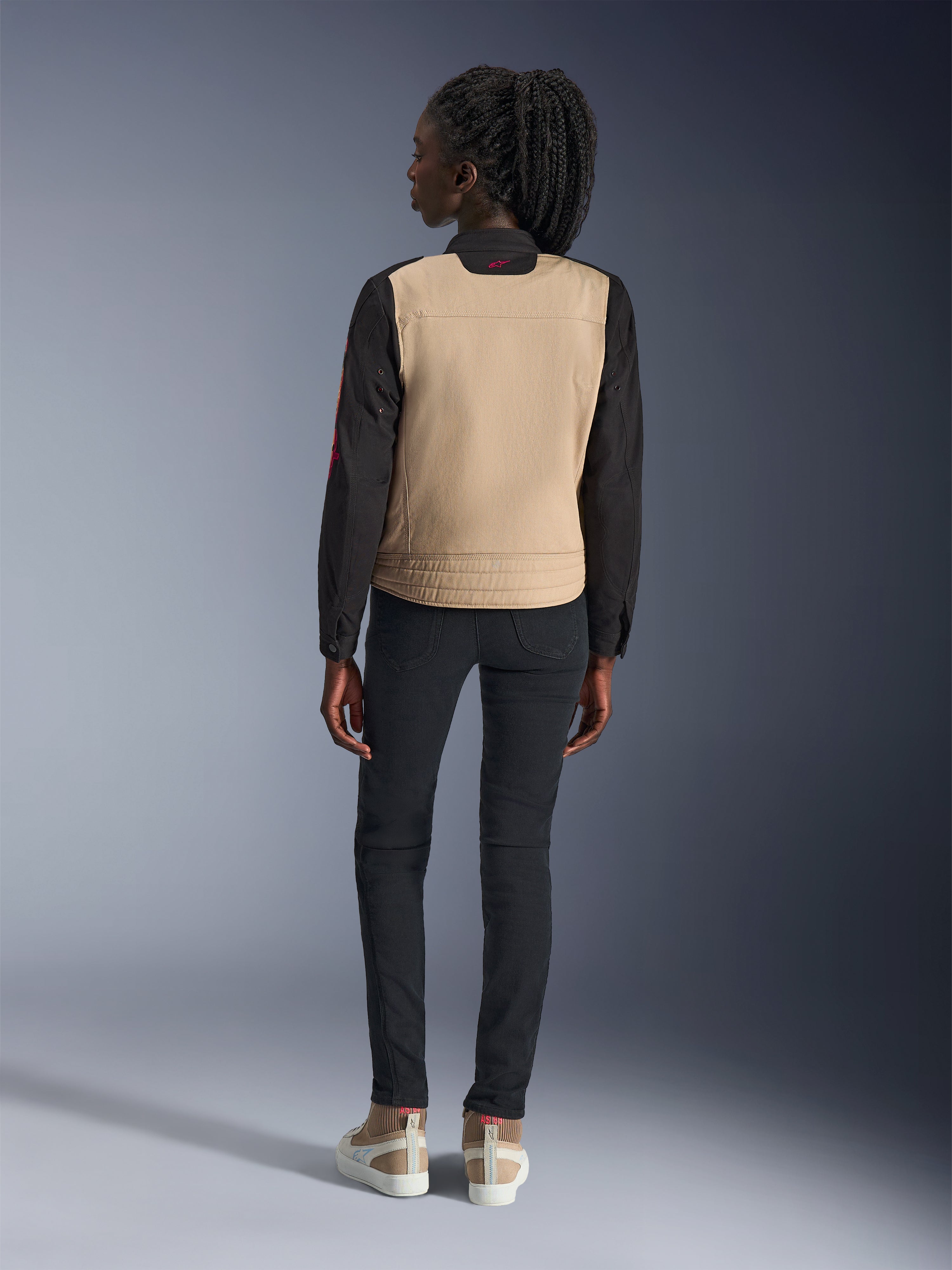 Femme portant la veste Alpinestars Stella Grace, Aluminum Black avec torse beige et manches noires, vue de dos, associée à un pantalon slim noir et des baskets décontractées, équipement de moto urbain pour femme
