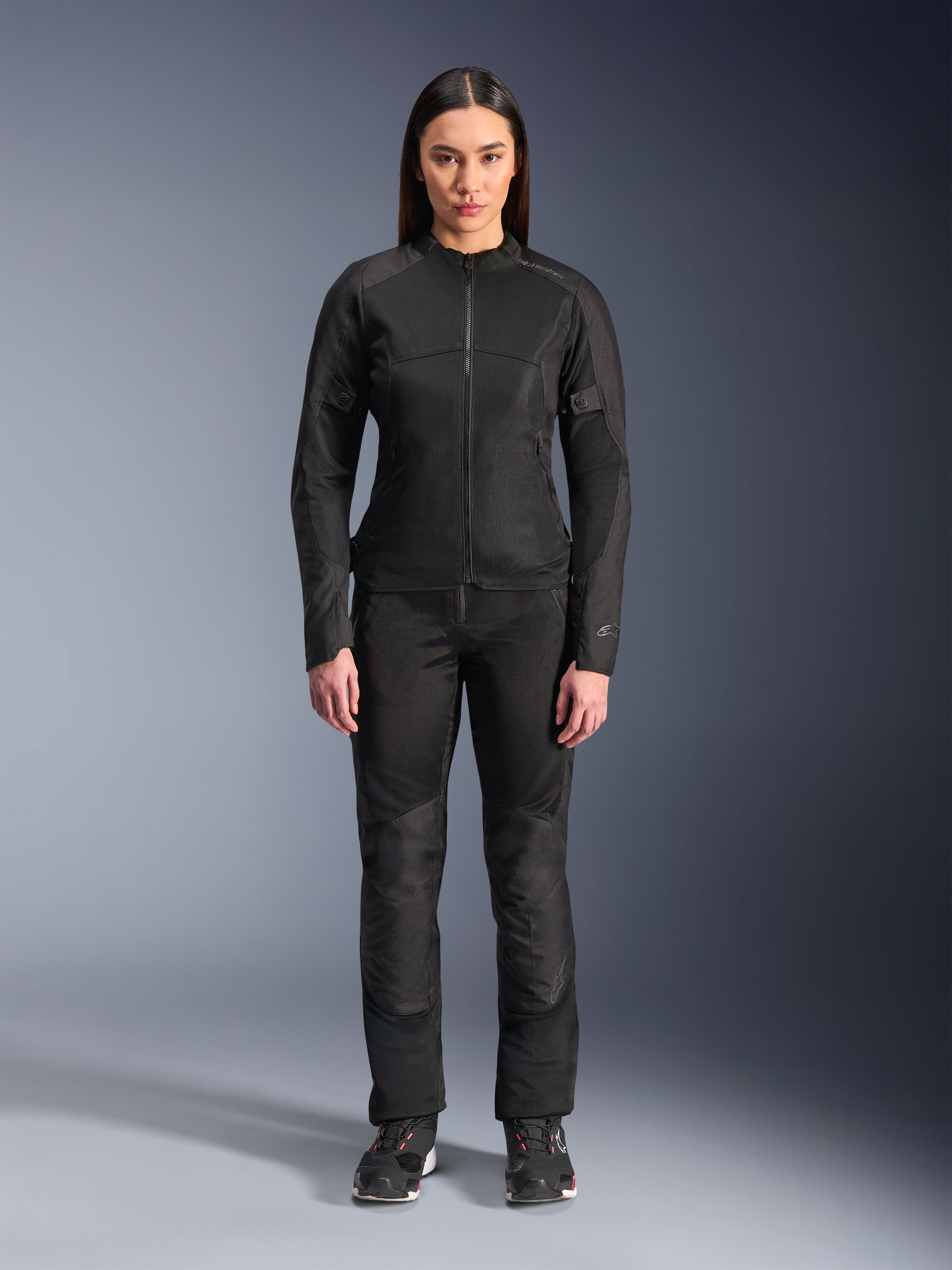 Frau trägt die Alpinestars Stella C-1 Air Jacke in Schwarz, kombiniert mit passender schwarzer Textil-Motorradhose und Motorradschuhen, mit einem eleganten urbanen Design und großen Mesh-Einsätzen für Luftzirkulation.