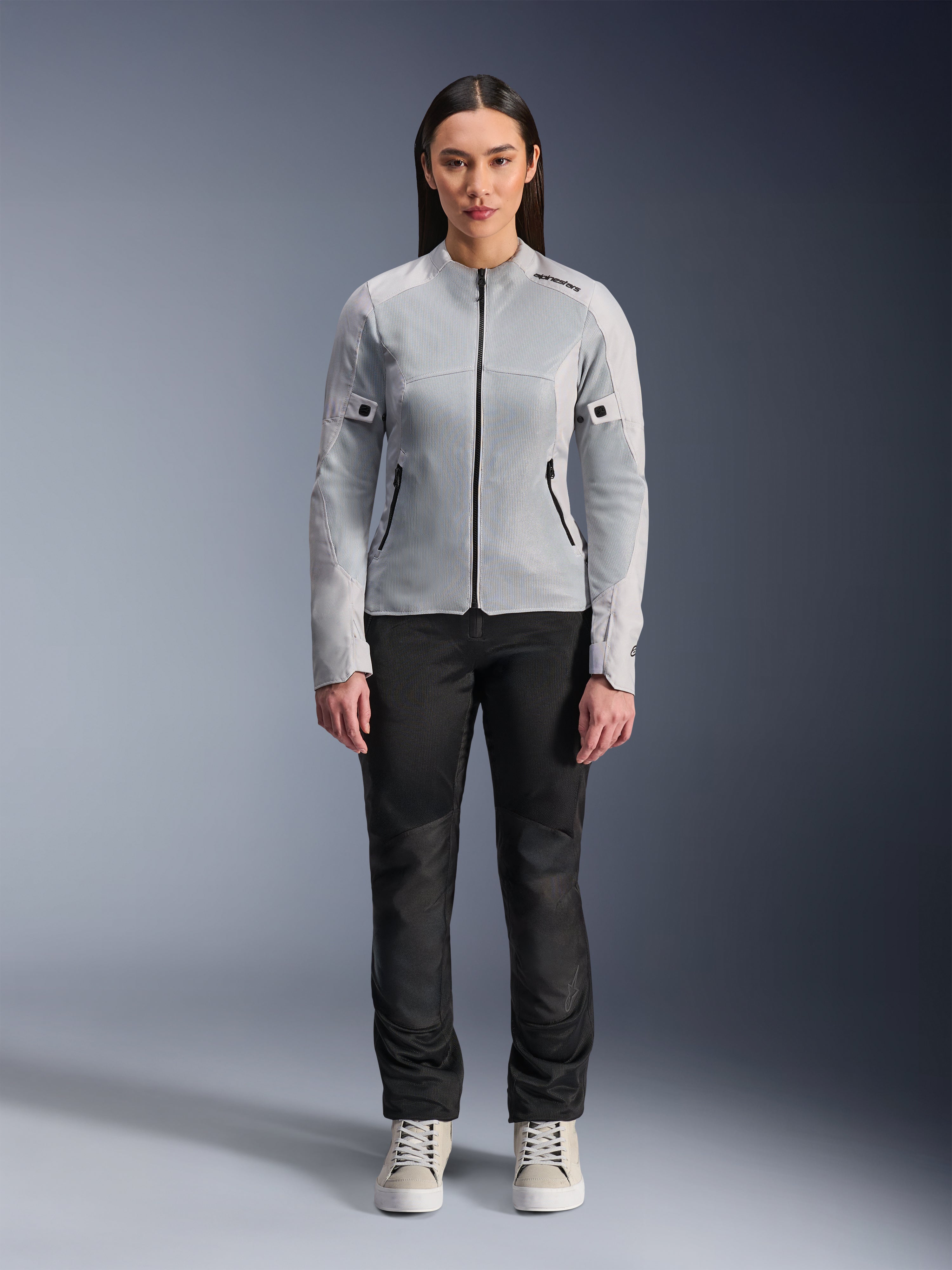 Femme portant la veste Alpinestars Stella C-1 Air en coloris argent, vue de face, veste de moto urbaine en textile avec panneaux en maille technique, associée à un pantalon noir et des baskets blanches, équipement de moto contemporain