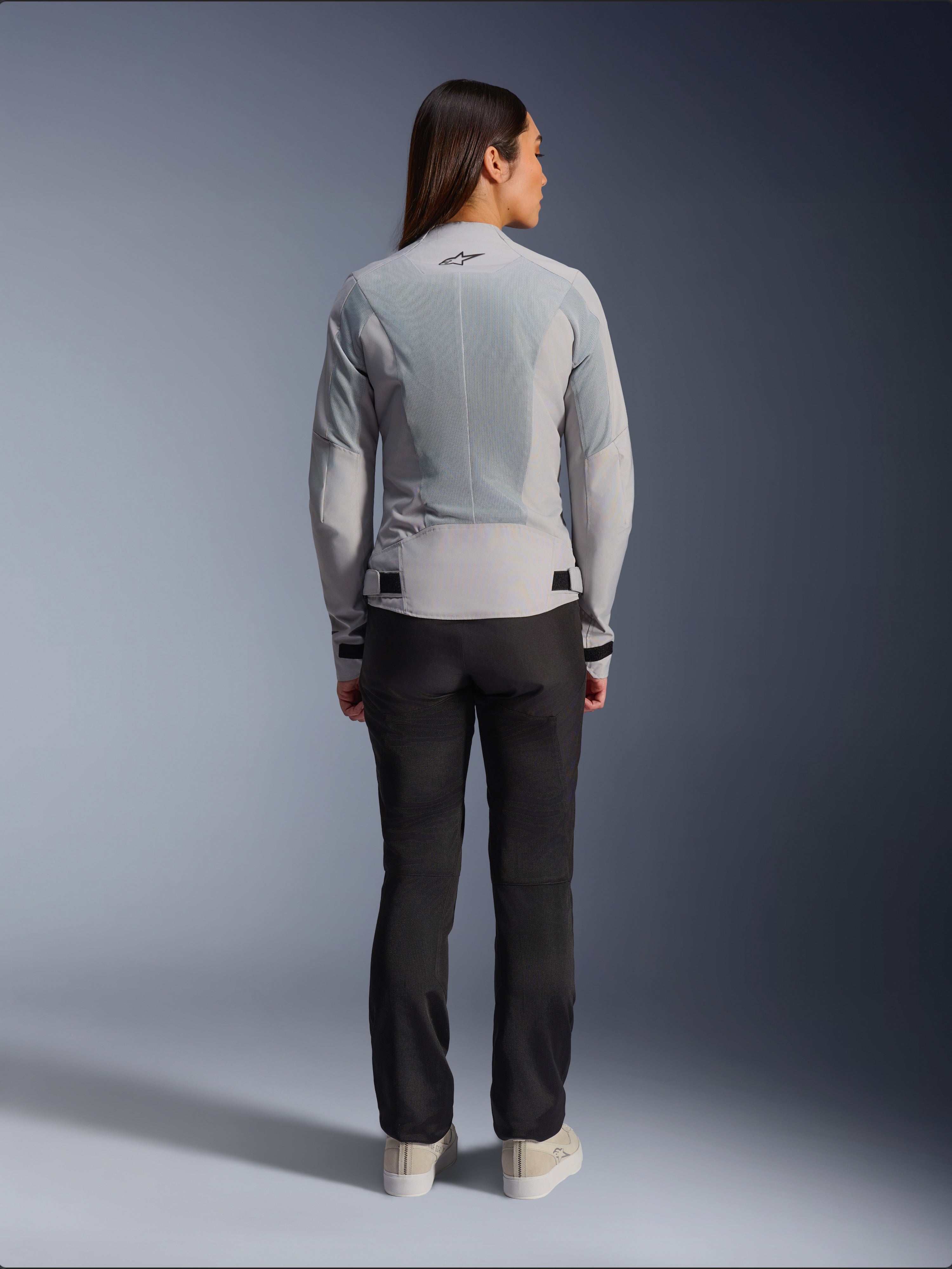 Femme portant la veste Alpinestars Stella C-1 Air couleur argent, vue de dos, dotée de grands panneaux en maille technique pour la ventilation et d'un logo étoile réfléchissant, associée à un pantalon de moto noir et des baskets blanches, équipement de moto urbain contemporain