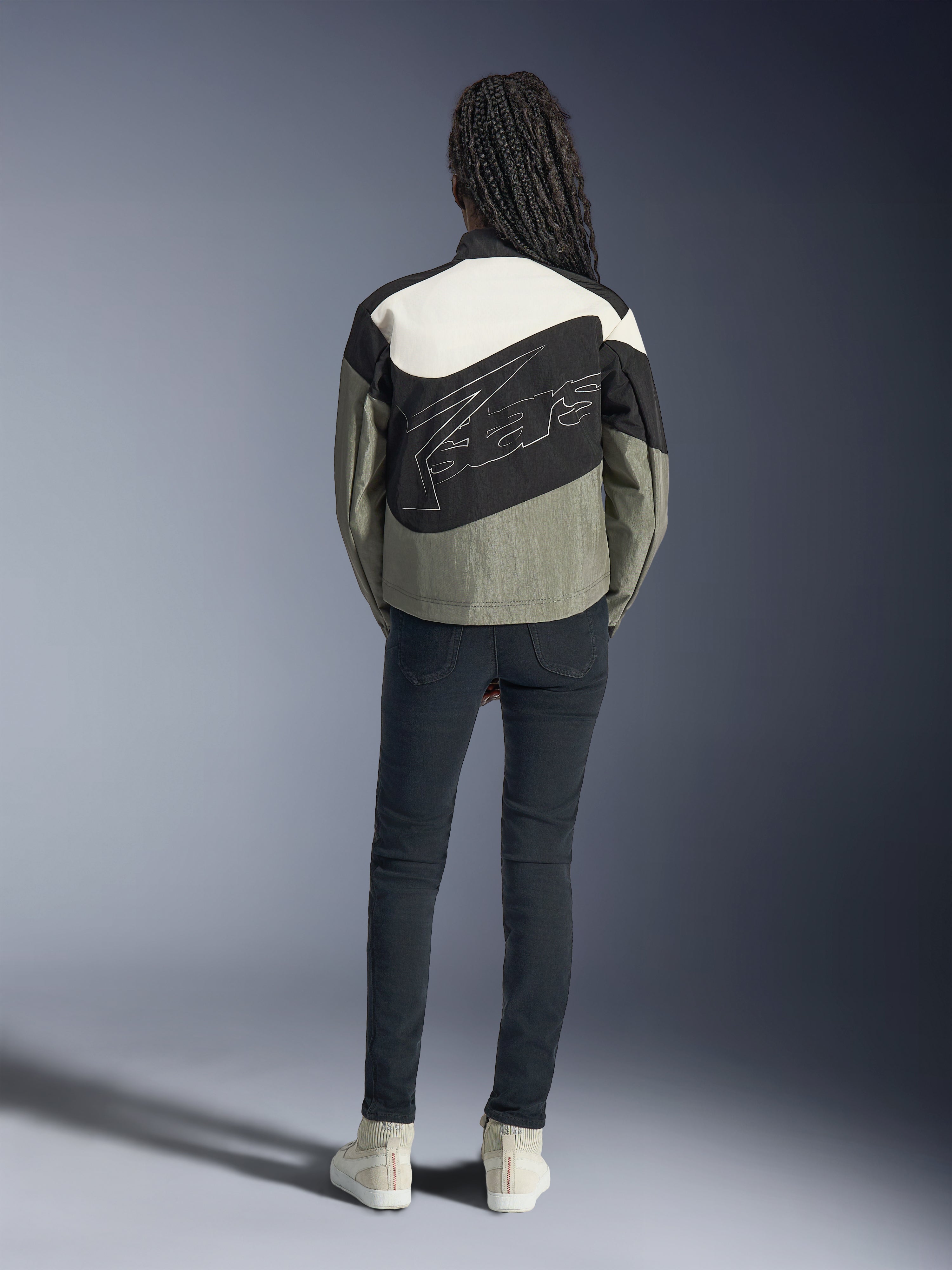 Achteraanzicht van een model in de Alpinestars Stella Moshell Jas in zwart, grijs en gebroken wit, voorzien van een asymmetrisch ontwerp en groot gestileerd logo op de rug, gecombineerd met een donkere slim-fit jeans en witte sneakers, op urban streetwear geïnspireerde motorkleding voor dames
