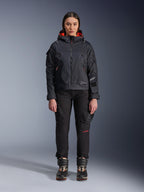 Mannequin femme portant la veste Alpinestars Molly Air en Noir et Corail Fluo, design d'inspiration utilitaire avec capuche et doublure corail, associée à un pantalon de moto noir et des bottes de moto, équipement de moto urbain contemporain