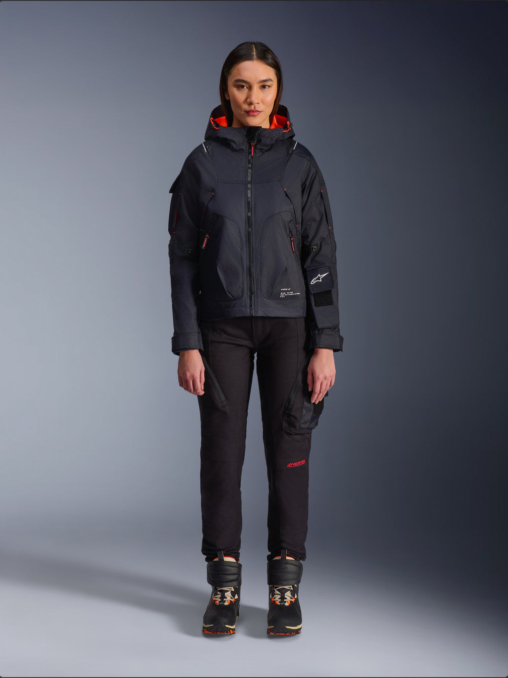 Alpinestars Actea damesbroek, zwarte motorbroek met rode tekstdetails en cargozakken, gedragen door een vrouwelijke motorrijder met een zwart jack met capuchon, moderne urban motorkleding