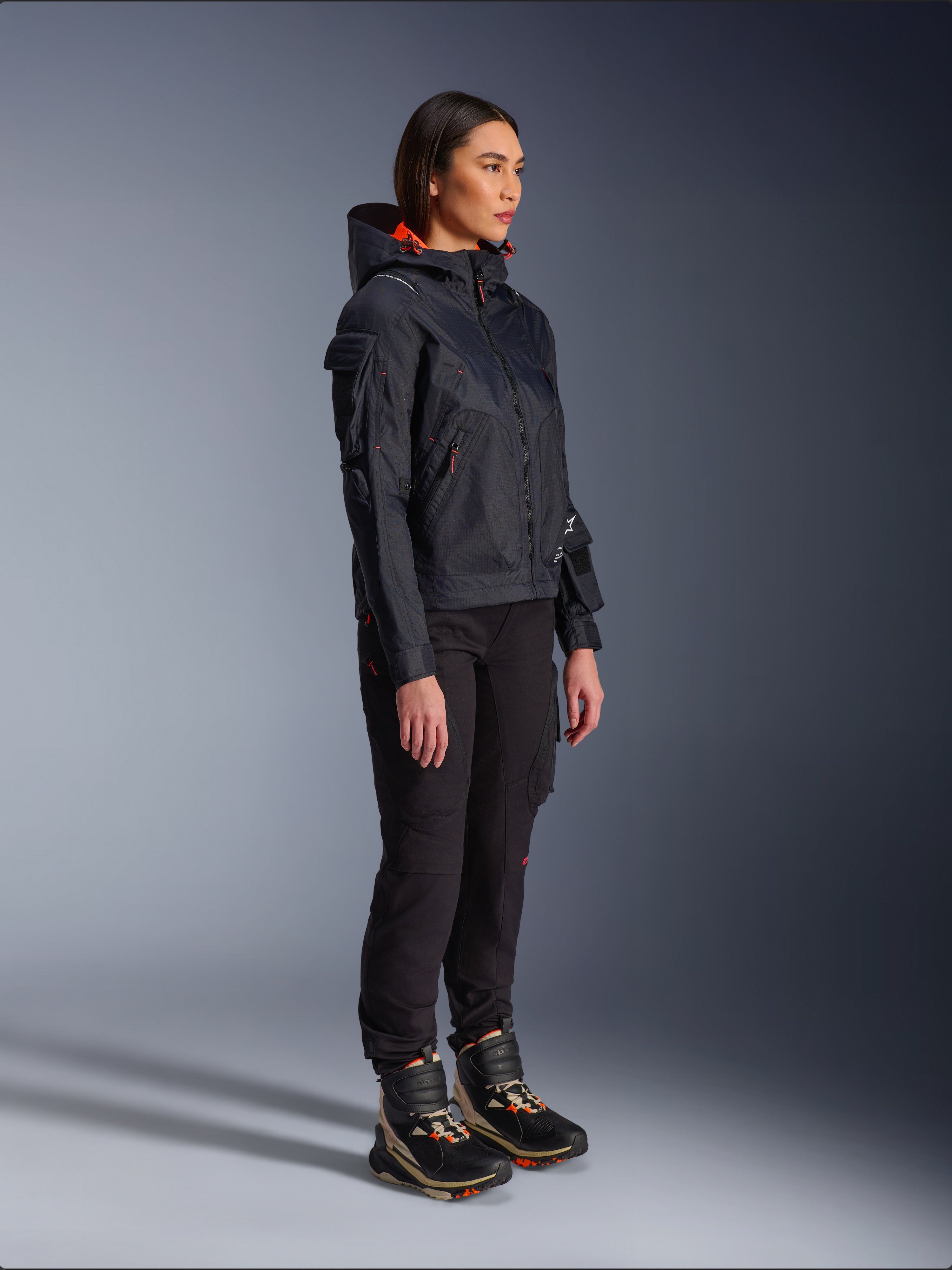Veste Alpinestars Molly Air en Noir Corail Fluo, veste de moto légère en textile avec doublure de capuche orange vif, portée par une motarde avec un pantalon de moto noir et des bottes techniques, design d'inspiration utilitaire urbain contemporain.