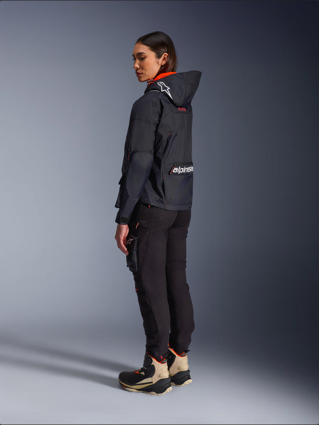 Motarde portant la veste Alpinestars Molly Air en Noir Corail Fluo, vue latérale arrière montrant la capuche avec logo Astars et grand marquage Alpinestars au dos, associée à un pantalon cargo de moto noir et des bottes d'aventure beiges et noires, équipement urbain contemporain pour femmes