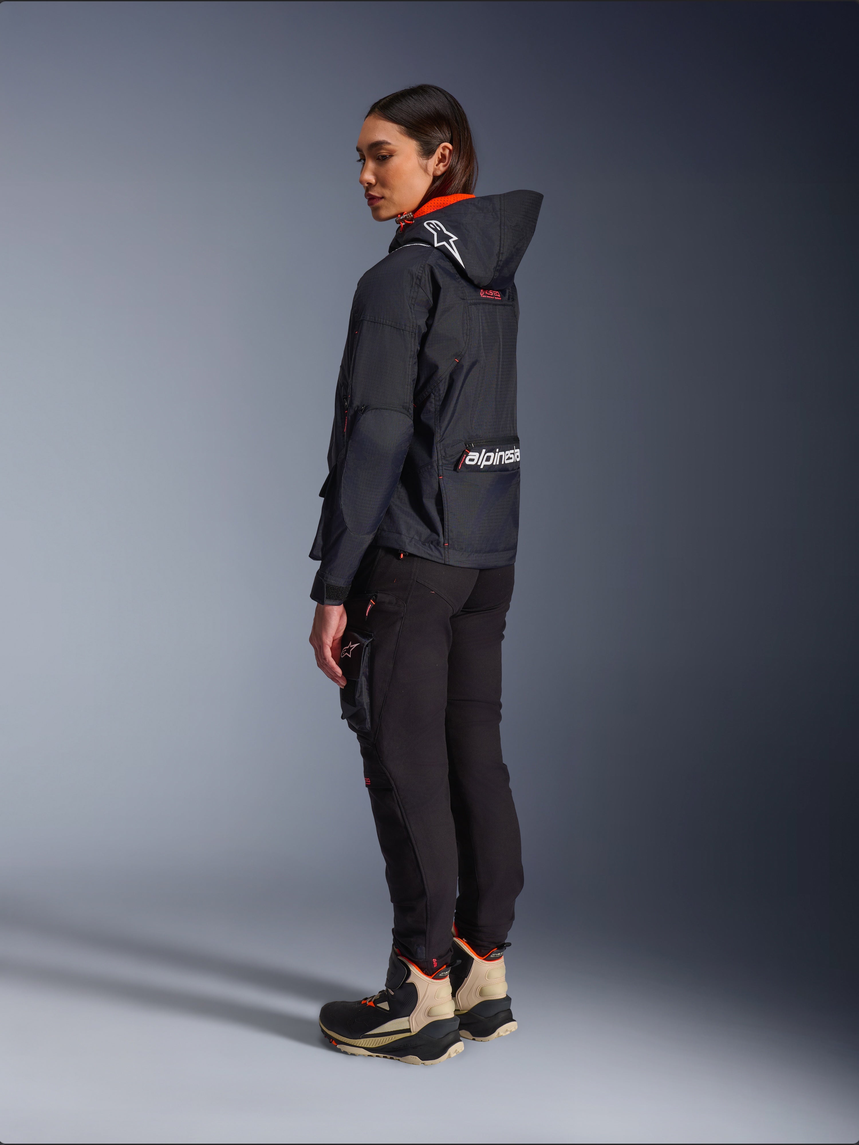 Motarde portant la veste Alpinestars Molly Air en Noir Corail Fluo, vue latérale arrière montrant la capuche avec logo Astars et grand marquage Alpinestars au dos, associée à un pantalon cargo de moto noir et des bottes d'aventure beiges et noires, équipement urbain contemporain pour femmes