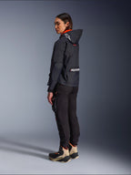 Alpinestars Actea dames motorbroek, zwart, moderne motorbroek met een taps toelopende pasvorm en cargo-zak op het bovenbeen, getoond vanuit een zij-achterperspectief op een model met een donker jack met capuchon en motorlaarzen, urban rijstijl.