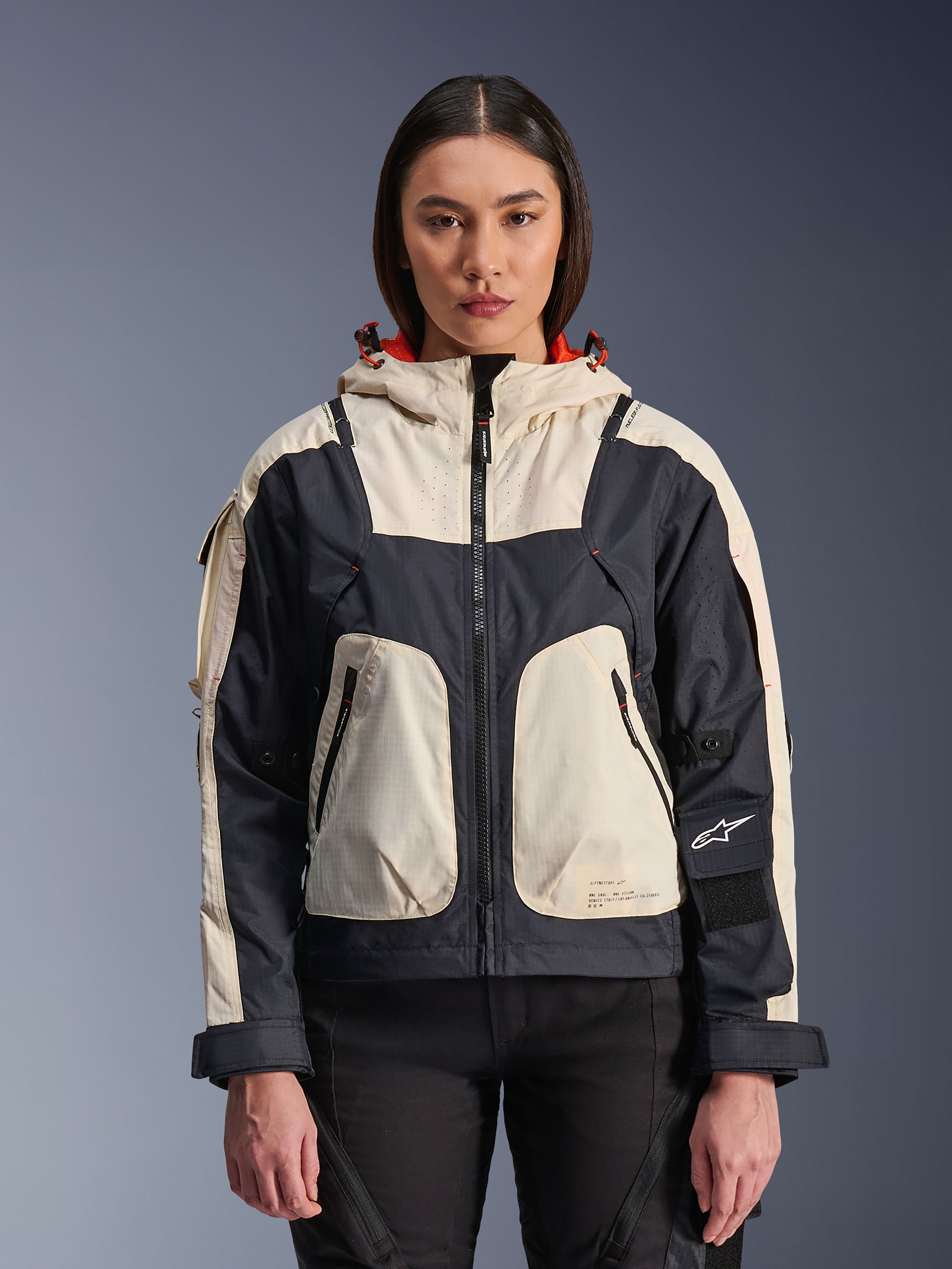 Alpinestars Molly Air Jacket, urban motorjas voor dames, Black Almond, vooraanzicht op een model, voorzien van crème en zwarte ripstop-panelen, capuchon met oranje voering, ritssluiting aan de voorzijde en grote utility-zakken, eigentijdse op streetwear geïnspireerde motorkleding