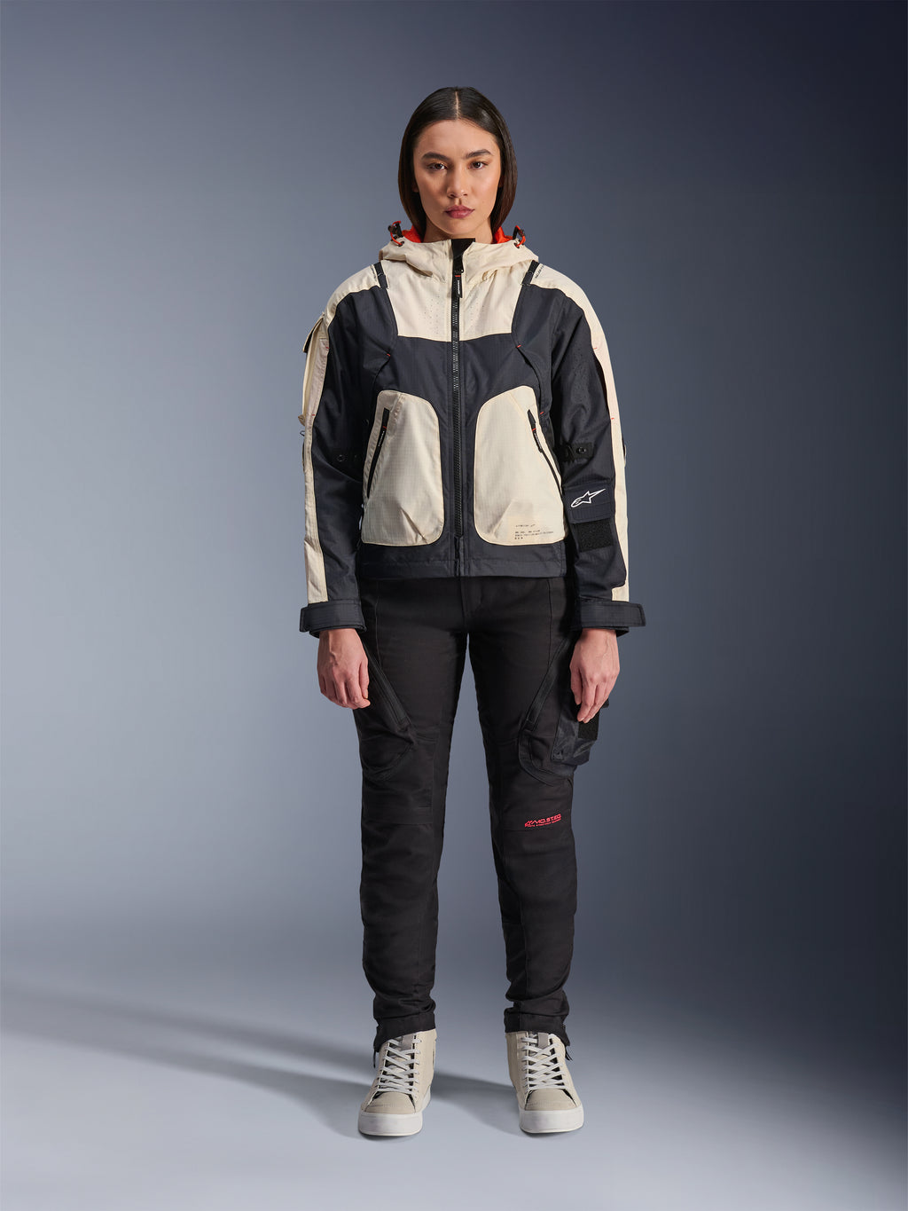 Vrouwelijk model draagt de Alpinestars Molly Air Jacket in Black Almond, voorzien van een color-blocked ontwerp met crème en zwarte panelen en een rood gevoerde capuchon, gecombineerd met een zwarte motorbroek en crème sneakers, studio-opname van stedelijke hedendaagse motorkleding.