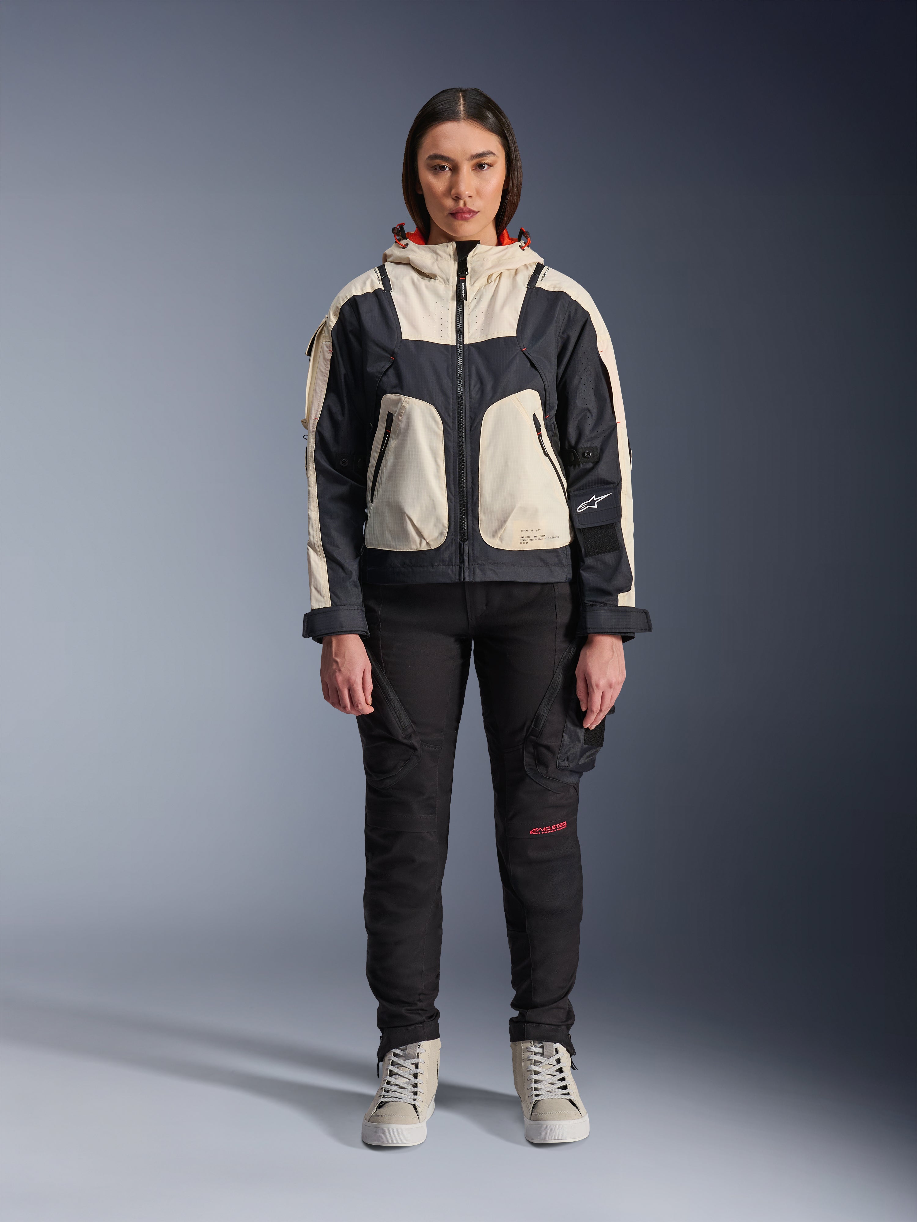 Vrouwelijk model draagt de Alpinestars Molly Air Jacket in Black Almond, voorzien van een color-blocked ontwerp met crème en zwarte panelen en een rood gevoerde capuchon, gecombineerd met een zwarte motorbroek en crème sneakers, studio-opname van stedelijke hedendaagse motorkleding.