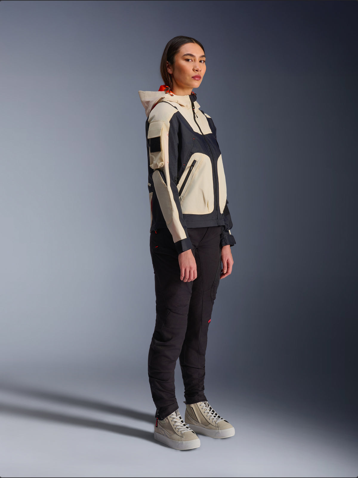 Alpinestars Molly Air Jacket in Black Almond, gedragen door een vrouwelijke rijder met zwarte broek en crèmekleurige hoge sneakers, voorzien van een tweekleurig ontwerp met zwarte en beige panelen, geïntegreerde capuchon en eigentijdse urban stijl.
