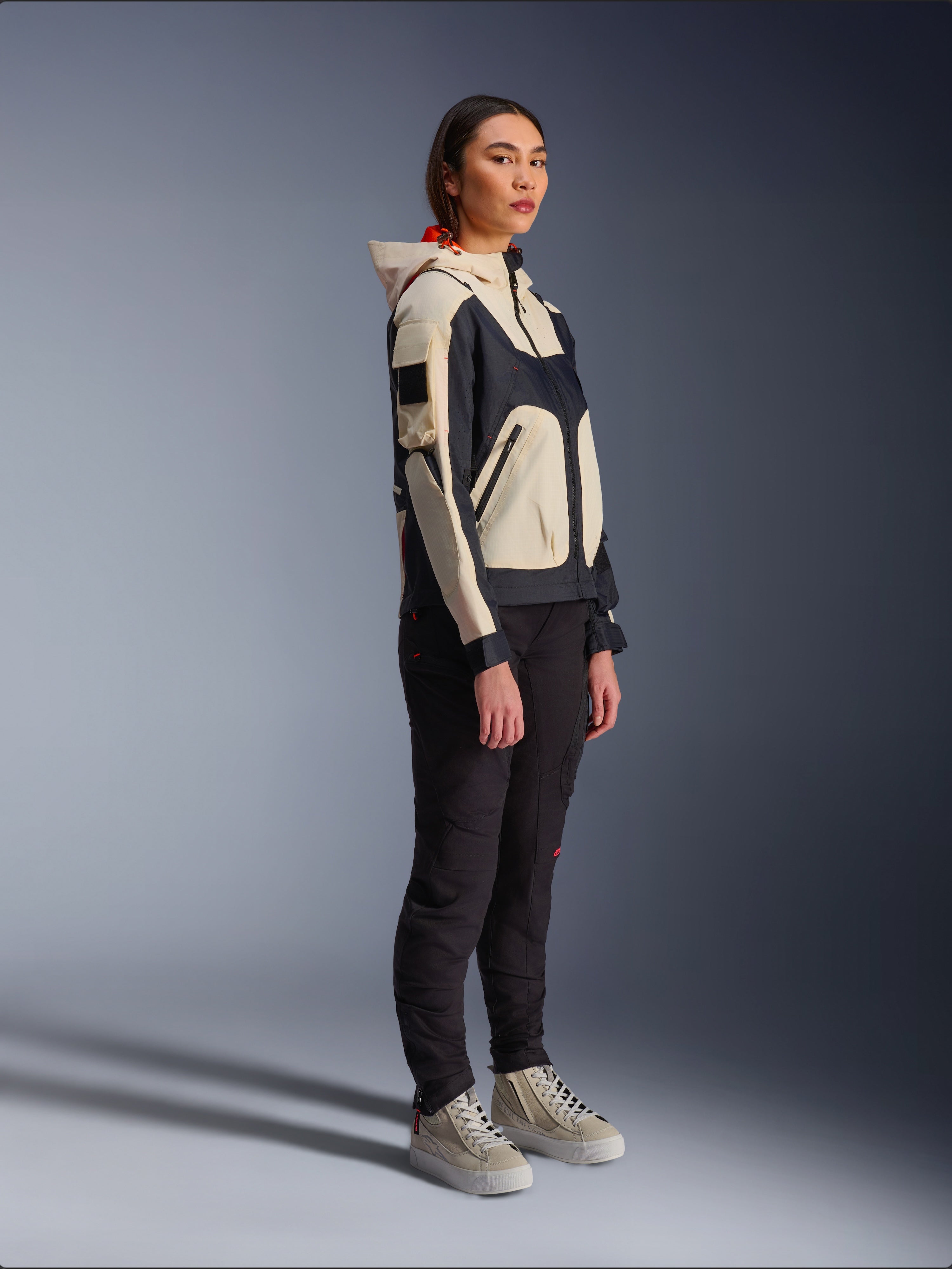 Alpinestars Molly Air Jacket in Black Almond, gedragen door een vrouwelijke rijder met zwarte broek en crèmekleurige hoge sneakers, voorzien van een tweekleurig ontwerp met zwarte en beige panelen, geïntegreerde capuchon en eigentijdse urban stijl.