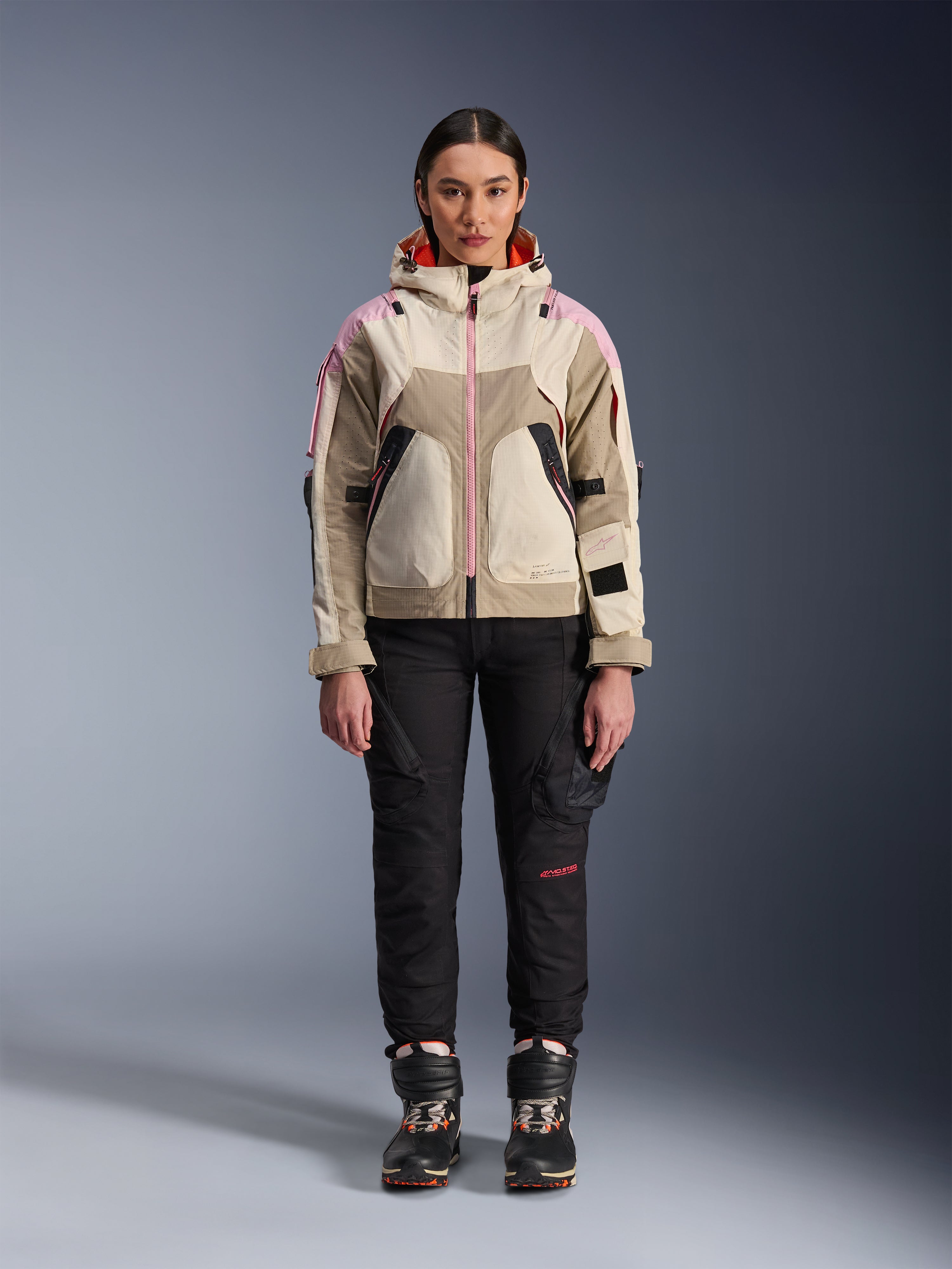 Vrouwelijk model draagt de Alpinestars Molly Air Jas in Sand Pink Almond, gecombineerd met een zwarte technische motorbroek en laarzen, vooraanzicht van het hele lichaam, urbane motorjas met utility-geïnspireerd ontwerp en roze accenten
