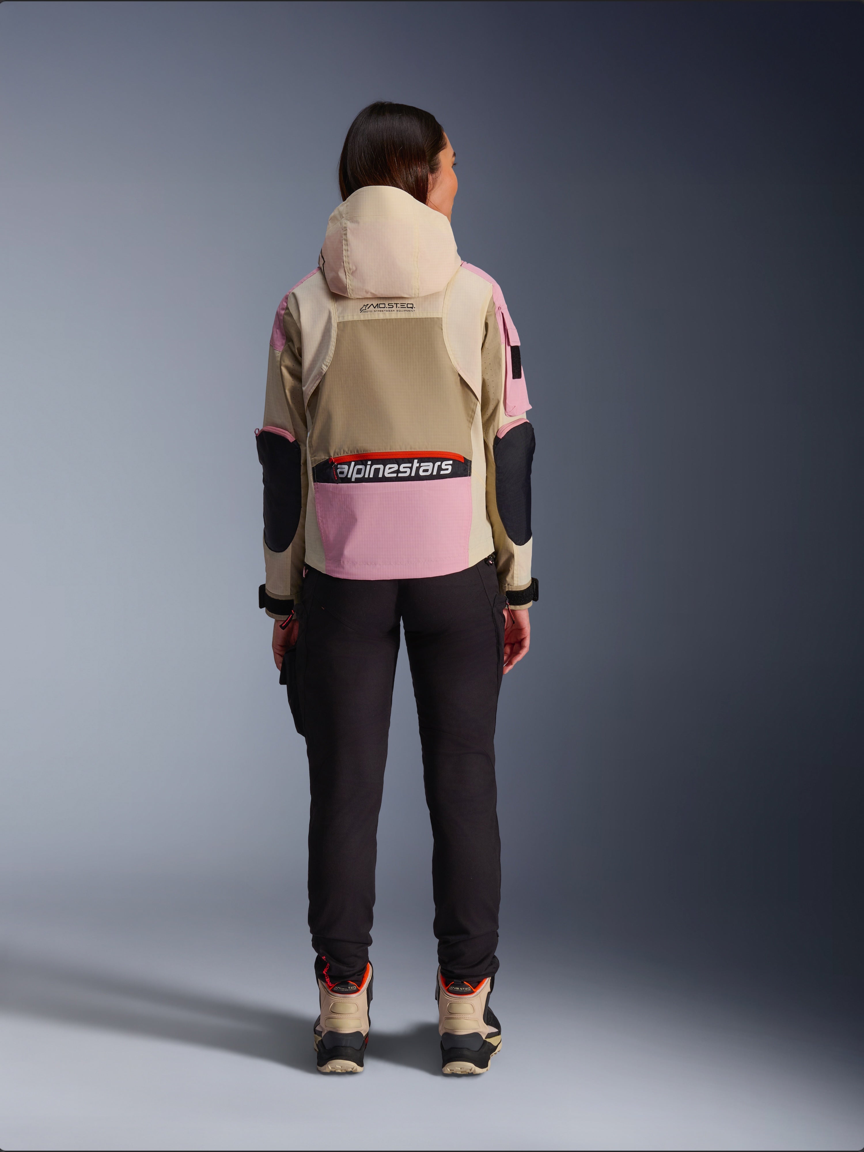 Vrouwelijke motorrijder van achteren gezien die de Alpinestars Molly Air Jacket draagt in Sand Pink Almond, met een zandkleurige capuchon en roze panelen met zwarte accenten, gecombineerd met een zwarte motorbroek en zandkleurige motorlaarzen, urban hedendaagse motorkleding