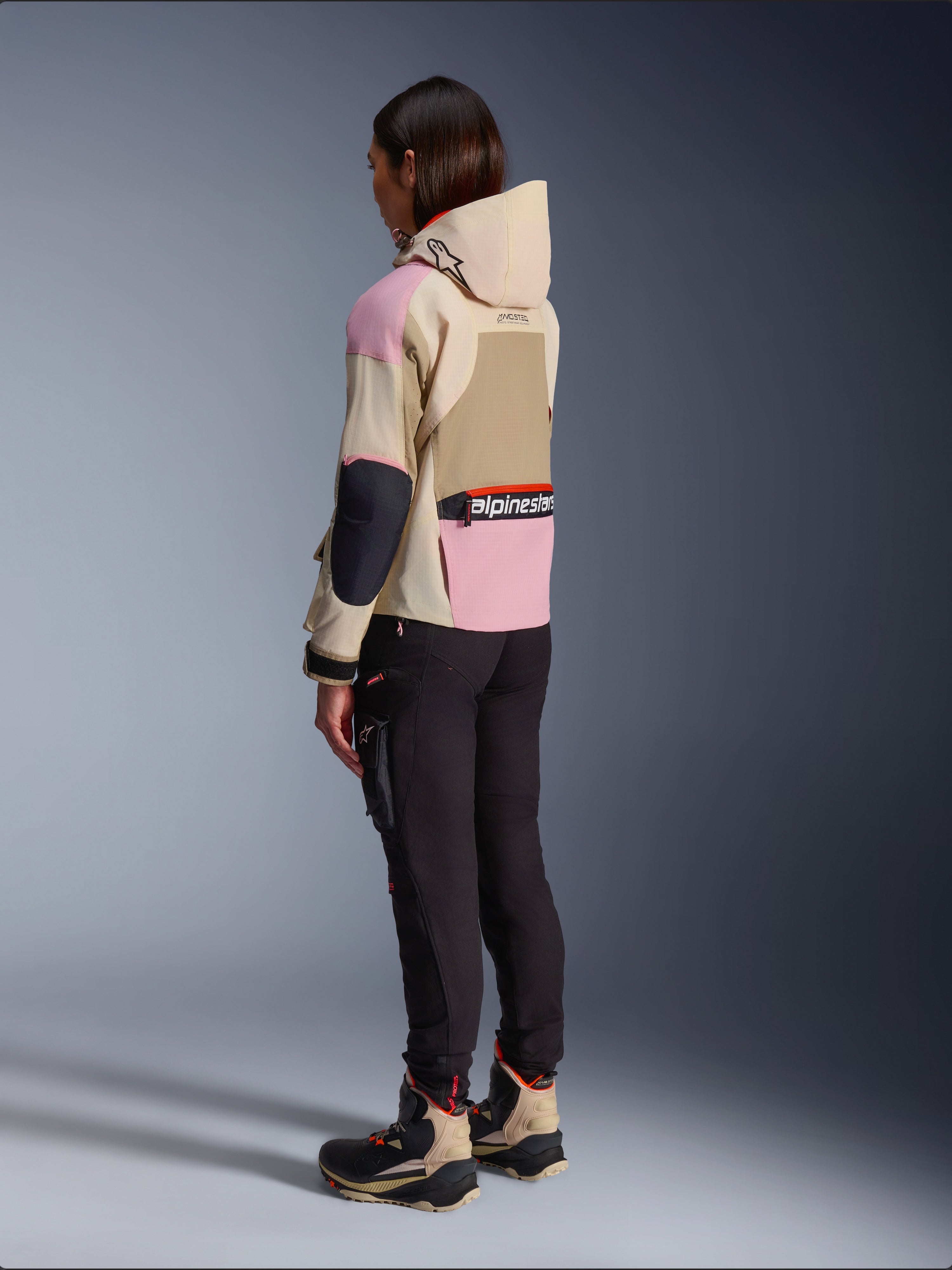 Vrouwelijke motorrijder die de Alpinestars Molly Air Jacket draagt in Sand Pink Almond, met een meerkleurig beige en roze ontwerp, achteraanzicht van opzij met zwarte motorbroek en bijpassende technische laarzen, urban motorstijl met capuchon.
