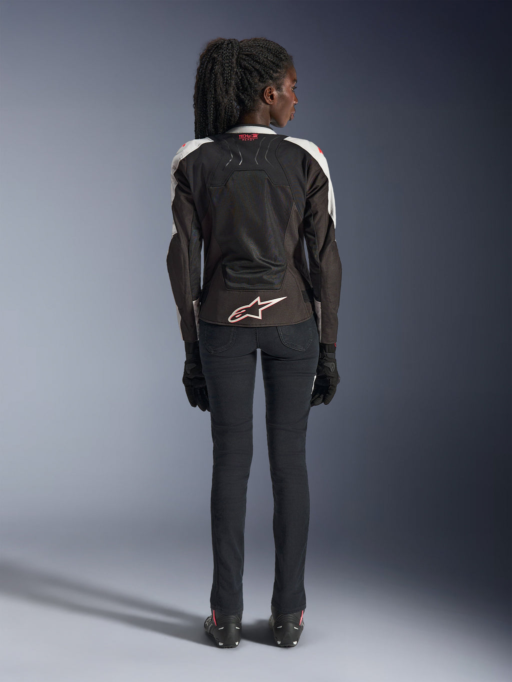 Veste Alpinestars Stella SMX Air pour femme, Ice Gray Black Coral Fluo, vue arrière montrant les panneaux en mesh ventilé, logo Alpinestars blanc au bas du dos et marquage Tech-Air Ready au col, portée avec un pantalon de moto noir et des gants.