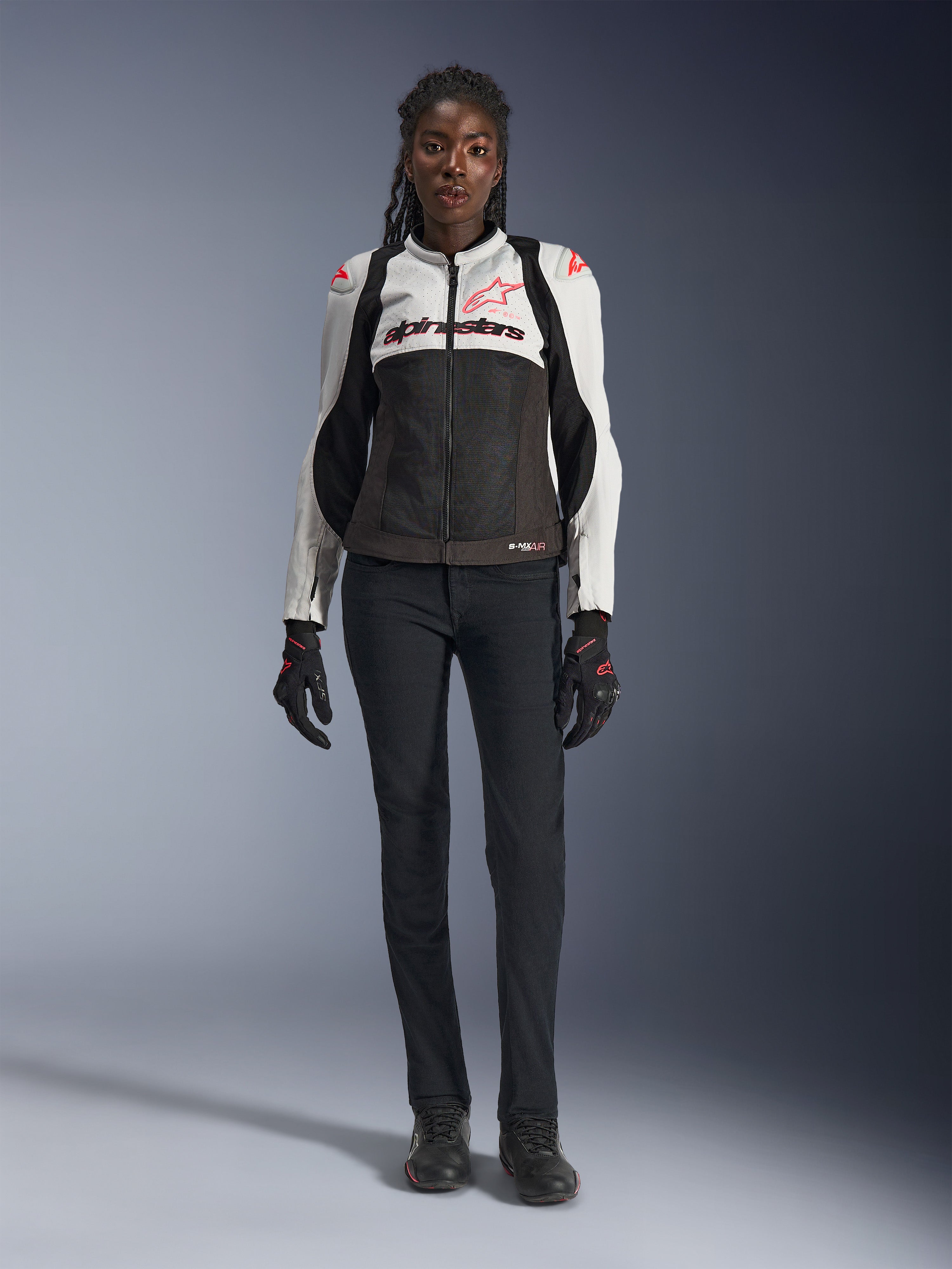 Veste Alpinestars Stella SMX Air, Ice Gray, noir et corail fluo, portée par un mannequin femme avec un pantalon de moto et des gants noirs, dotée d'un châssis en mesh et de sliders de protection aux épaules, équipement de moto de sport pour femme