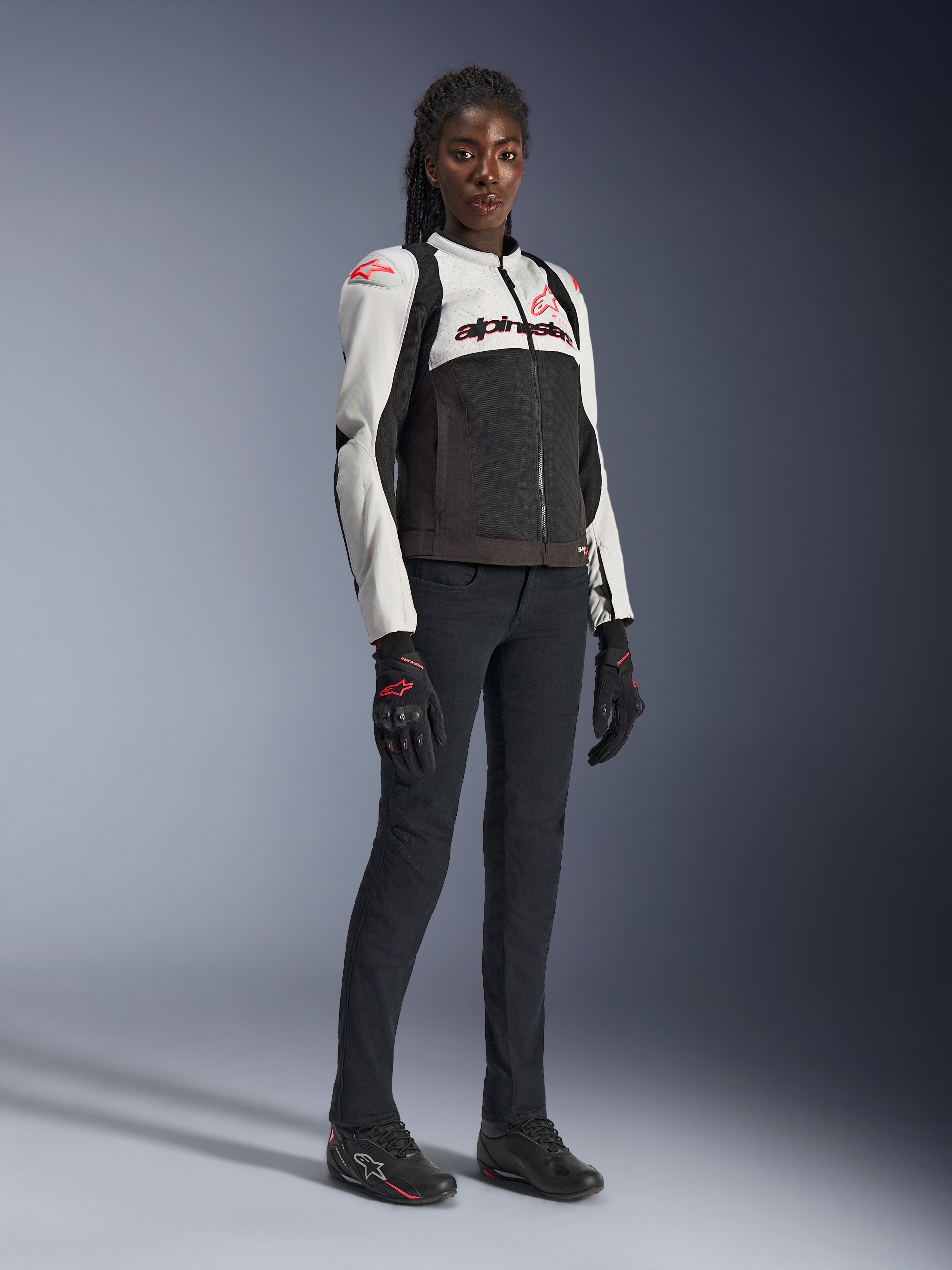 Veste Alpinestars Stella SMX Air pour femme, coloris Gris Glace, Noir et Corail Fluo, portée par un modèle féminin avec un pantalon et des gants de moto noirs, construction en mesh et logos rouge corail, équipement de moto sportif pour femme