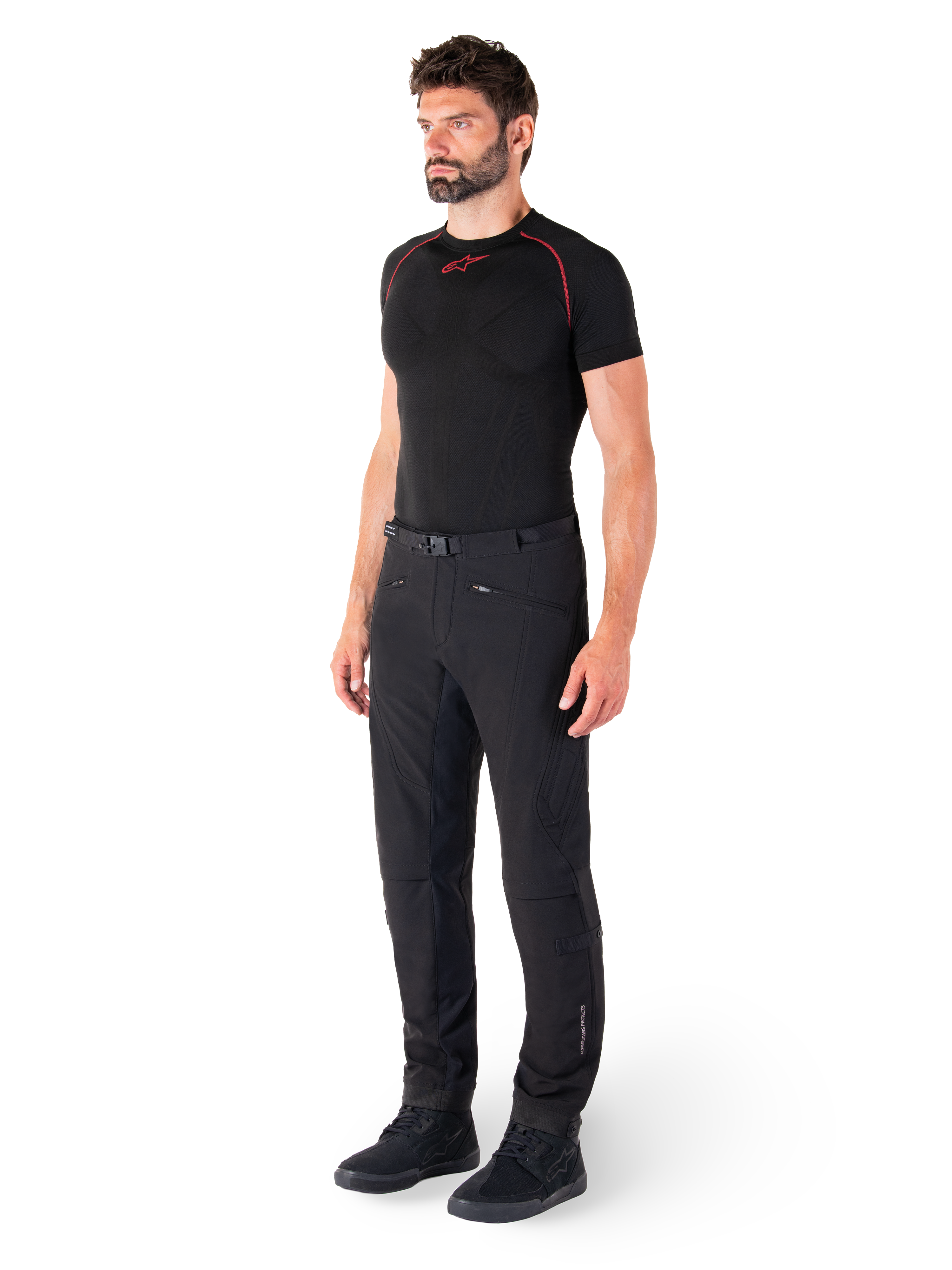 Pantalon Alpinestars Alden, pantalon de moto urbain, noir, textile softshell à coupe régulière, porté par un mannequin avec une couche de base noire et des chaussures de moto, doté d'une ceinture à fermeture magnétique et de poches zippées.