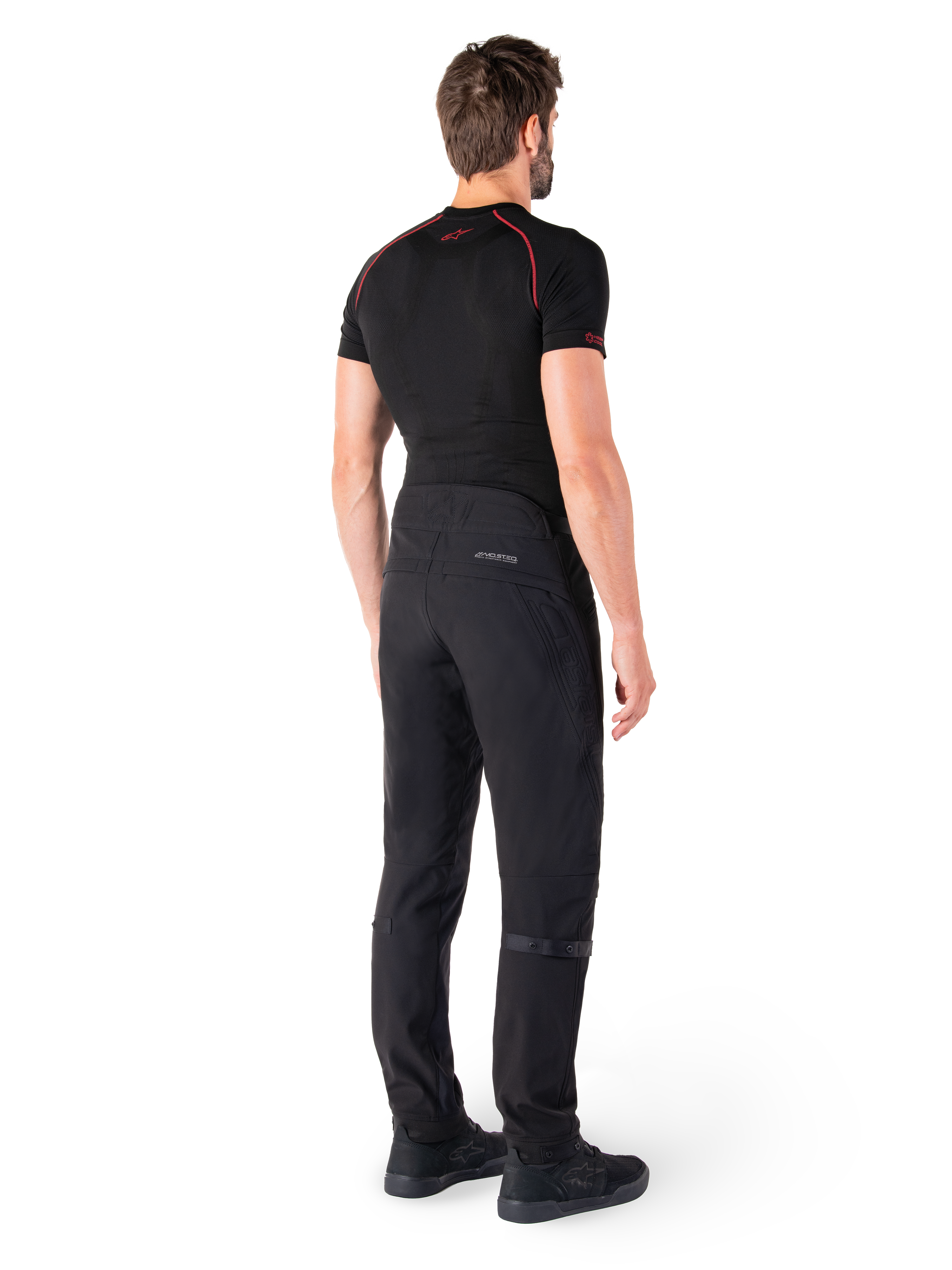 Pantalon Alpinestars Alden, noir, pantalon de moto urbain présenté de dos sur un mannequin, porté avec une couche de base noire à manches courtes et des chaussures de moto noires, avec des détails matelassés et le marquage réfléchissant MO.ST.EQ. sur l'empiècement arrière.