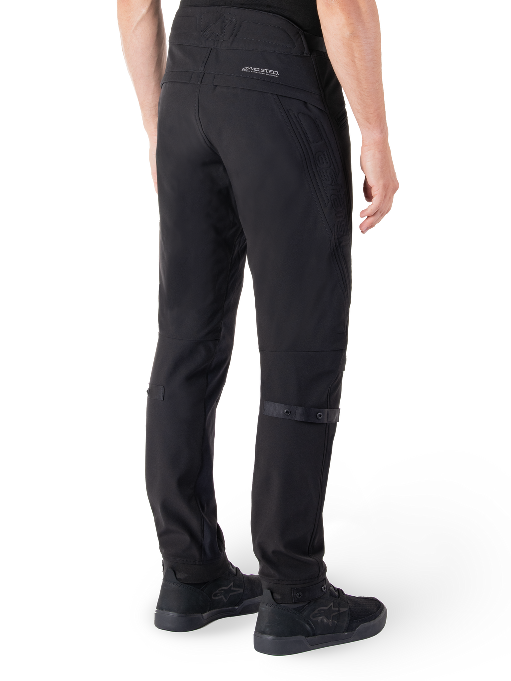 Pantalon Alpinestars Alden, pantalon de moto urbain, noir, vue arrière montrant le tissu softshell technique avec des détails matelassés, marquage réfléchissant MO.ST.EQ. sur l'empiècement arrière et sangles de mollet réglables, associé à des chaussures de moto noires
