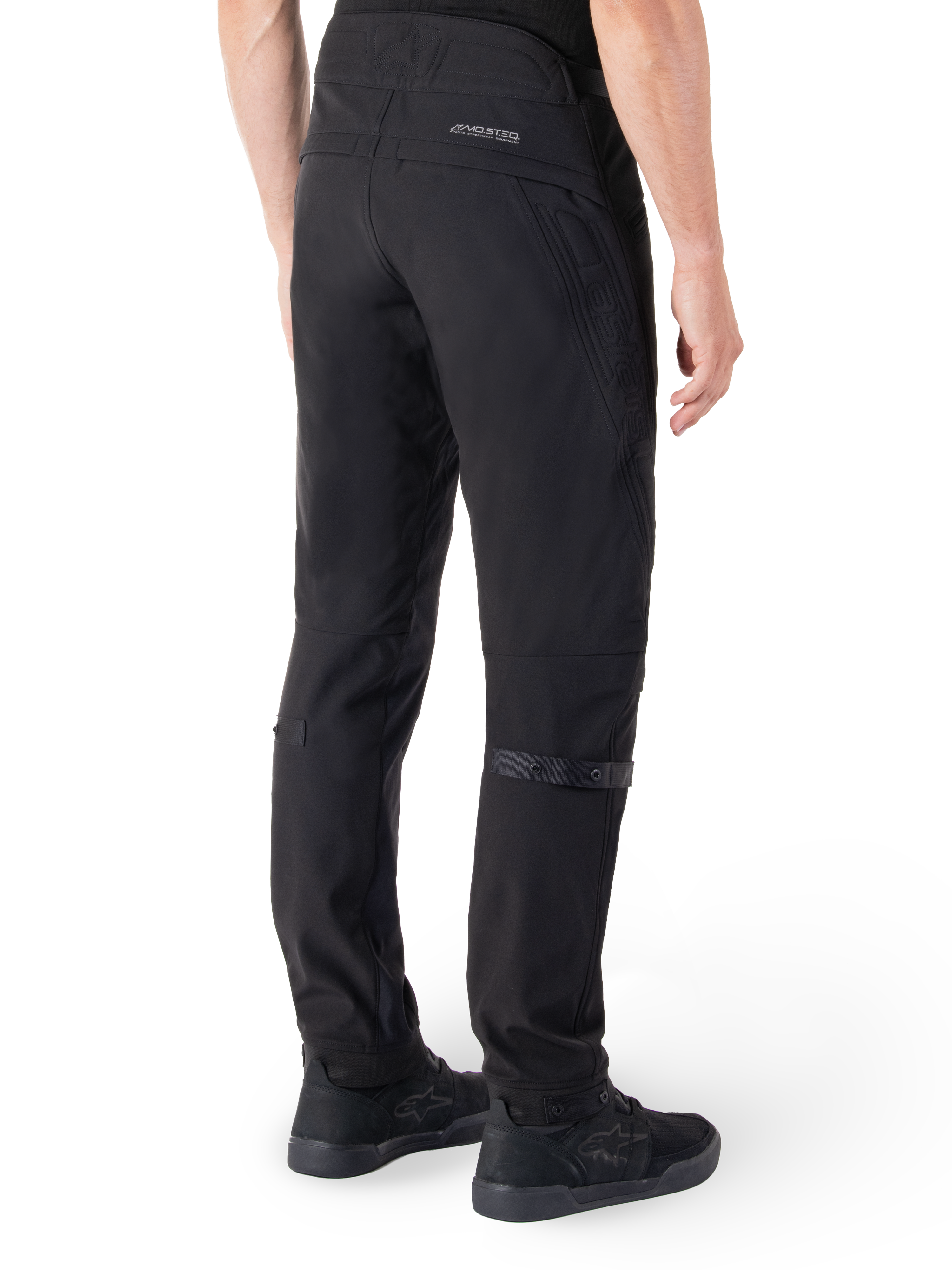 Pantalon Alpinestars Alden, pantalon de moto urbain, noir, vue arrière montrant le tissu softshell technique avec des détails matelassés, marquage réfléchissant MO.ST.EQ. sur l'empiècement arrière et sangles de mollet réglables, associé à des chaussures de moto noires