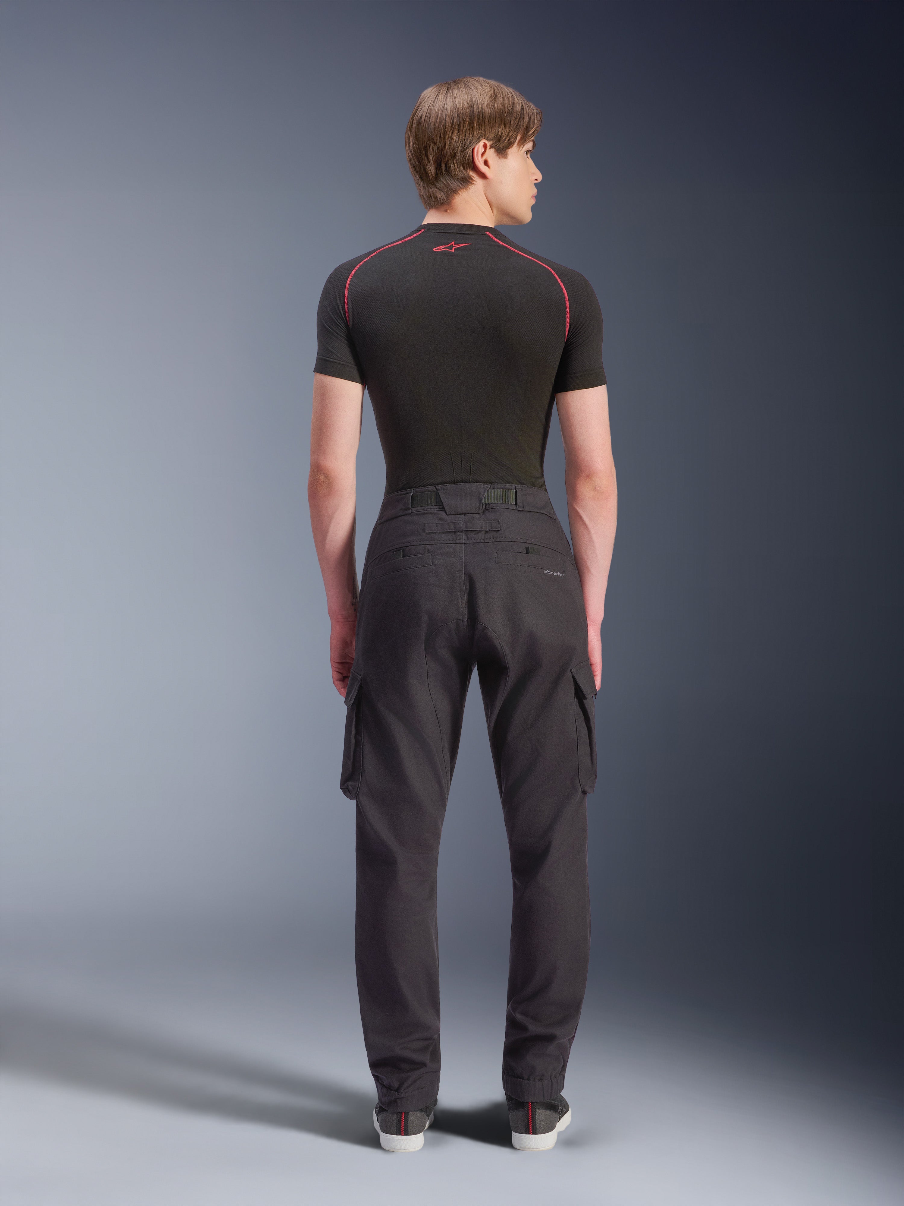 Alpinestars Flex-AST Cargo Pants - Short, zwarte stedelijke motorbroek van achteren gezien, met cargozakken aan de zijkant en elastische zomen, gedragen met een zwarte basislaag met korte mouwen en rode contraststiksels