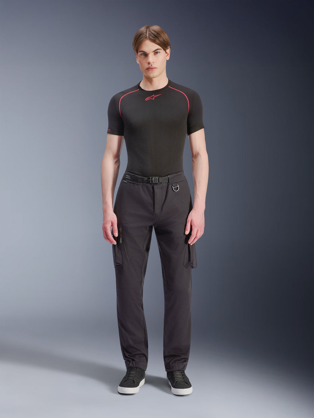 Alpinestars Flex-AST Cargo Broek - Short, zwart, gedragen door een mannelijk model met een zwart performance T-shirt en zwarte sneakers, stedelijke motorbroek met meerdere cargozakken, een taps toelopende pasvorm en een geïntegreerde riem met gesp en D-ring