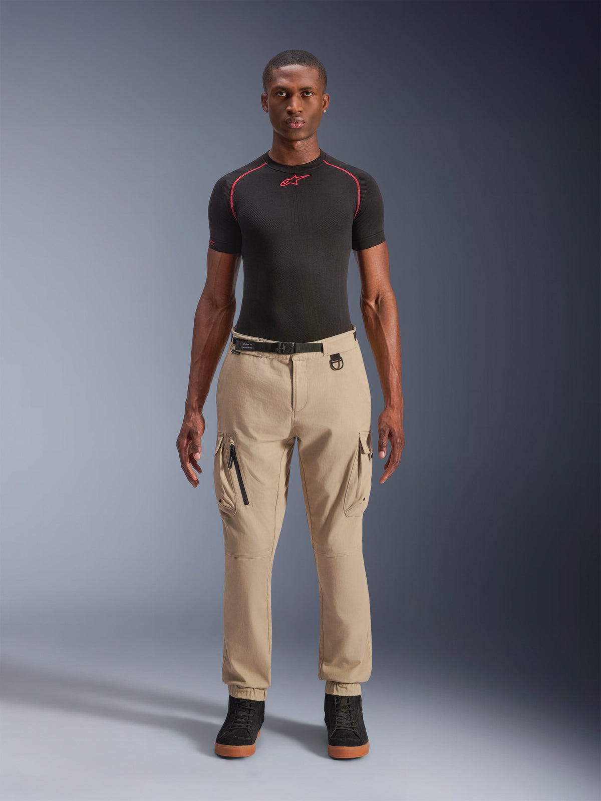 Alpinestars Flex-AST Cargo Hose - Short, Hellbeige urbane Motorradhose, reguläre konisch zulaufende Passform mit elastischen Säumen, funktionale Cargotaschen, integrierter schwarzer Gürtel mit Schnalle, getragen von einem männlichen Model mit schwarzem Base-Layer und Sneakern.