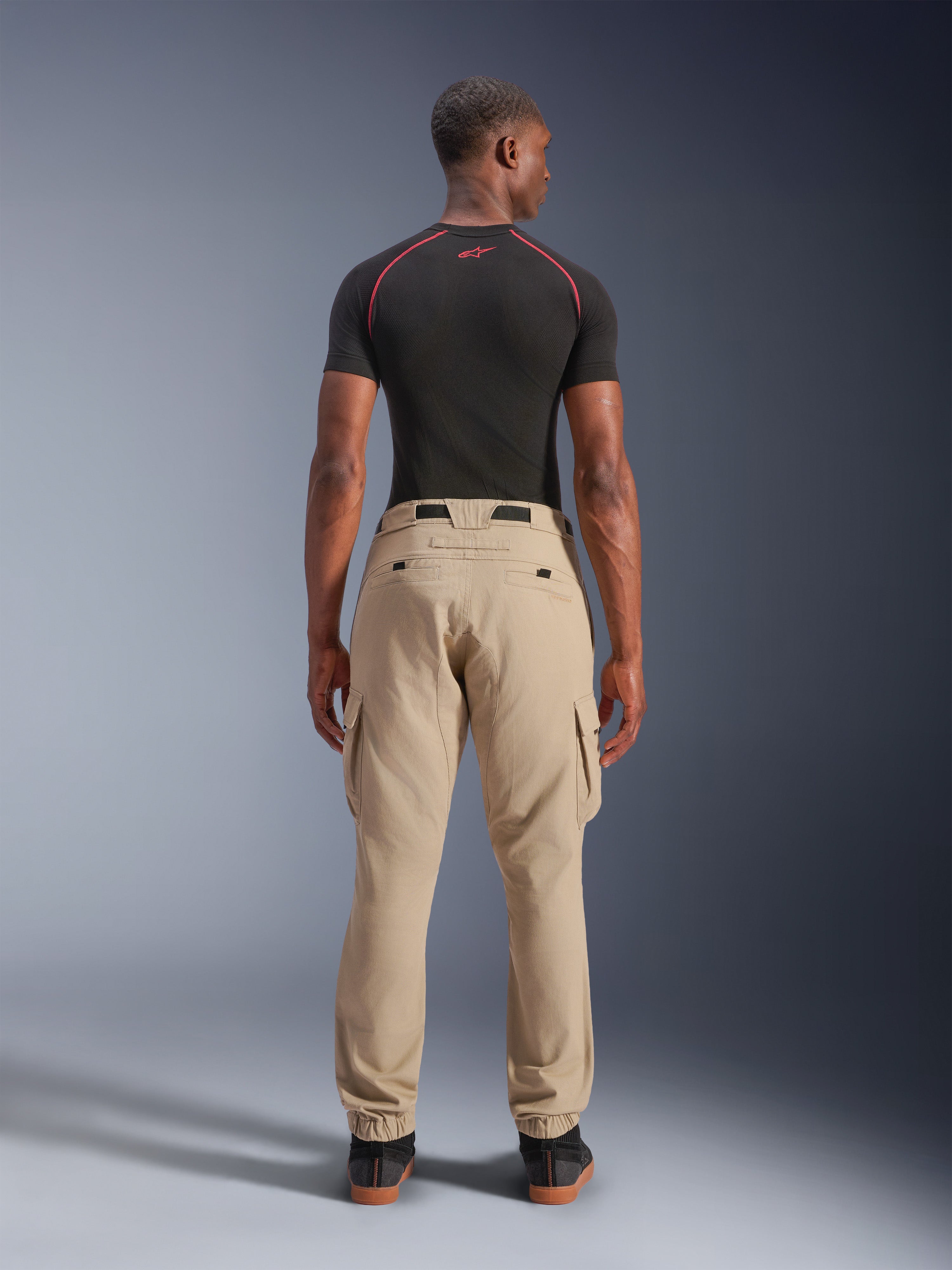 Alpinestars Flex-AST Cargo Pants, lichtbeige, achteraanzicht op een model, regular tapered pasvorm met cargozakken aan de zijkant en elastische zomen, gecombineerd met een zwarte basislaag met korte mouwen en rode accenten, stedelijke motorkleding