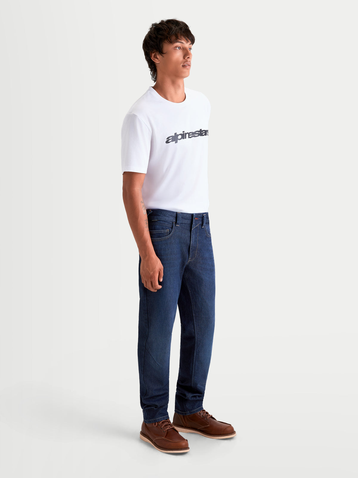 Alpinestars Flex-AST 7 Denim broek, heren motorjeans in donkerblauw, zijaanzicht op model met wit T-shirt en bruine laarzen, CE-gecertificeerde motorbroek van enkellaags denim met stretch