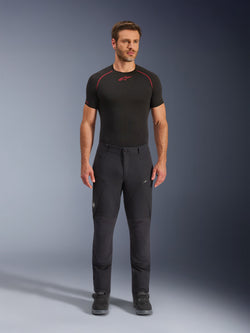 Alpinestars Flex-AST Explorer Pants, zwarte adventure touring motorbroek, man draagt nauwsluitende textiele rijbroek met cargozakken met rits en verstevigde panelen, gecombineerd met een zwarte basislaag en motorlaarzen, duurzame bescherming voor adventure rijden
